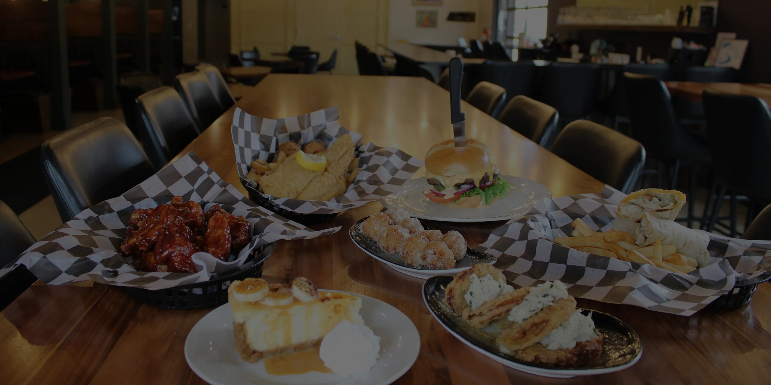 A variety of dishes displayed on a wooden table in a dining area, including a burger, chicken wings, and desserts.