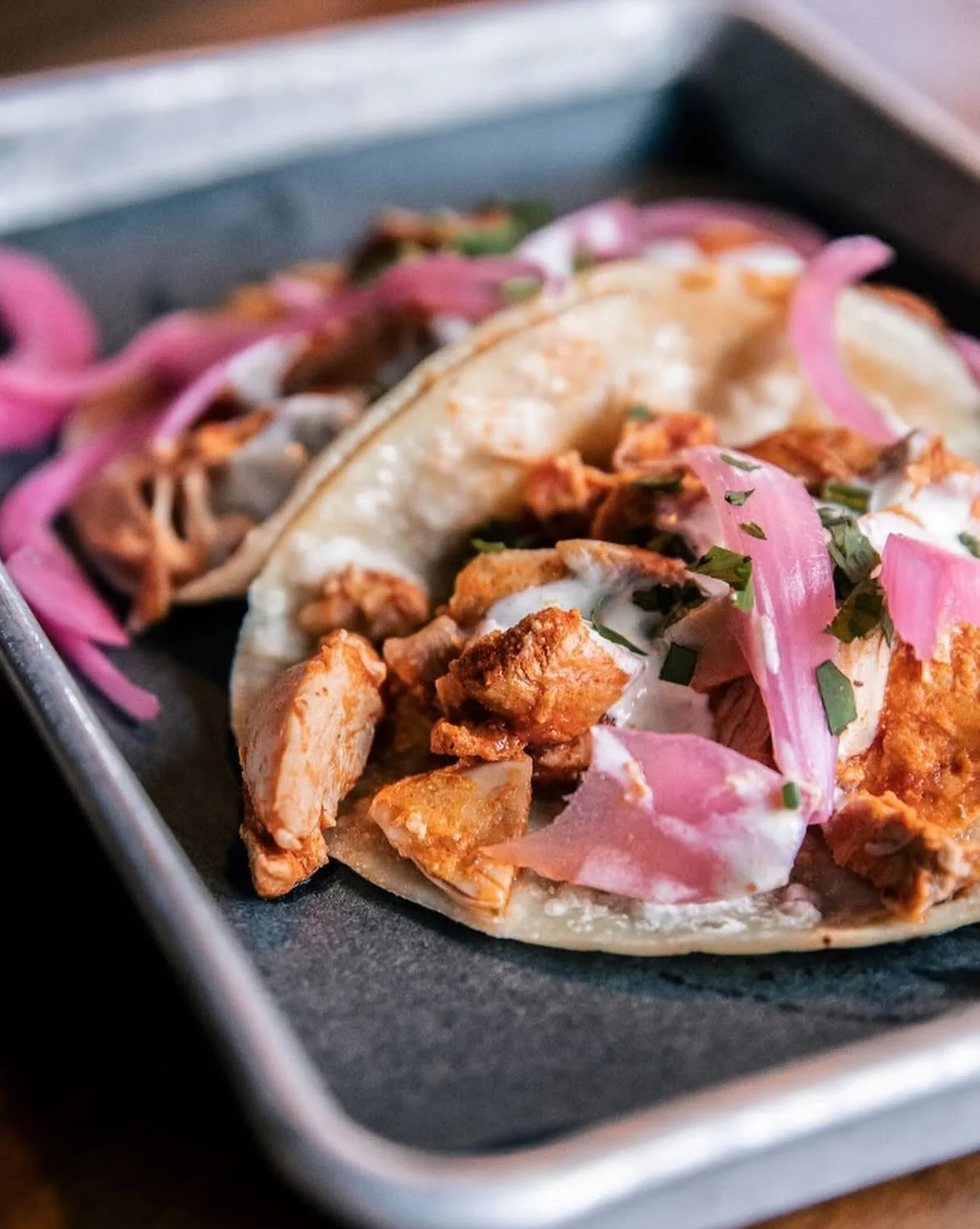 A close-up of tacos served on a tray, featuring soft tortillas filled with spiced meat, toppings of onions, cilantro, and a drizzle of sauce.