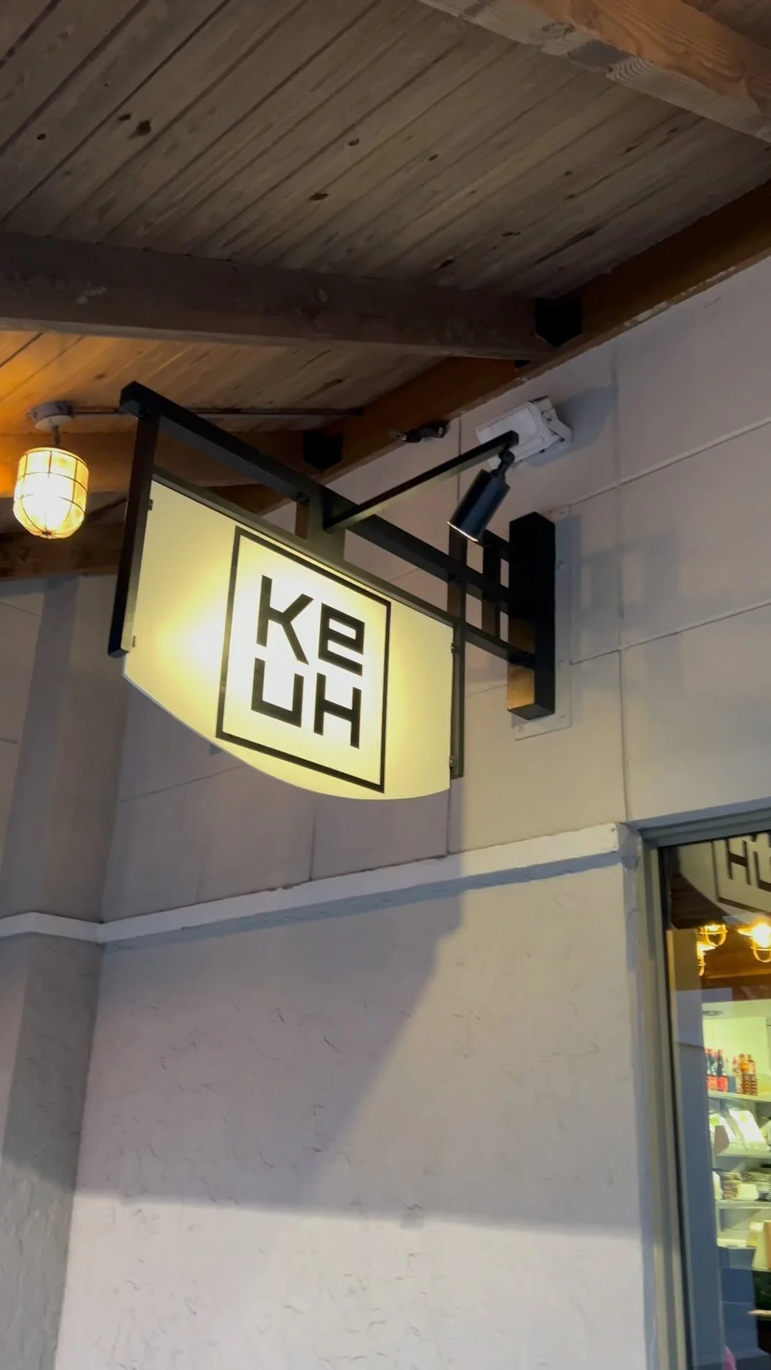 An illuminated exterior hanging sign for "KE-UH" restaurant with a minimalist black square logo, mounted on a white wall under a wooden overhang.