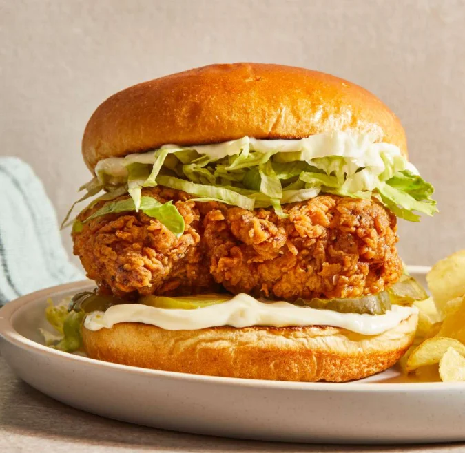 A crispy fried chicken sandwich with lettuce, mayonnaise, and pickles, served on a toasted bun with a side of potato chips.