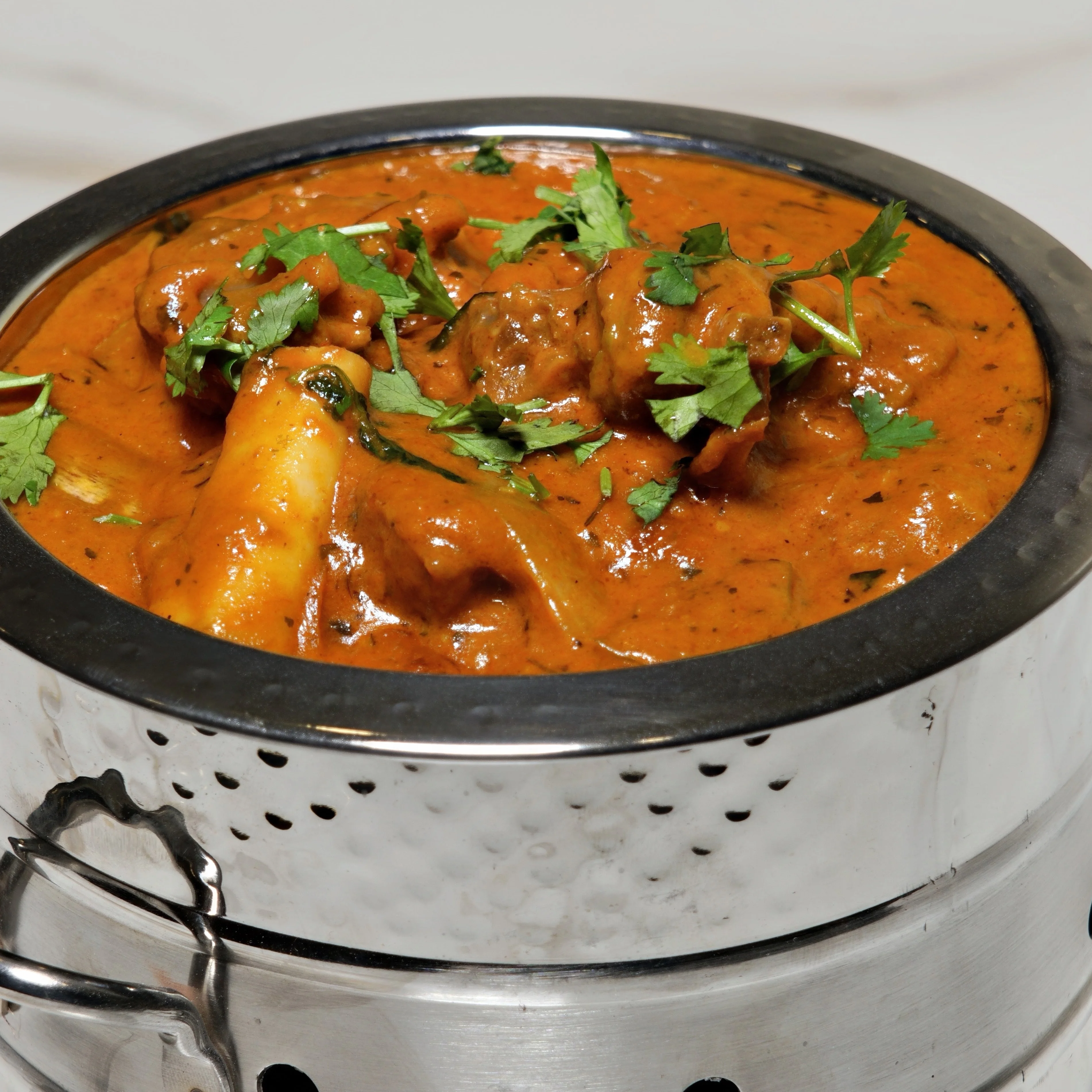 A rich and spicy curry featuring meat pieces, garnished with fresh cilantro, displayed in a traditional silver pot.
