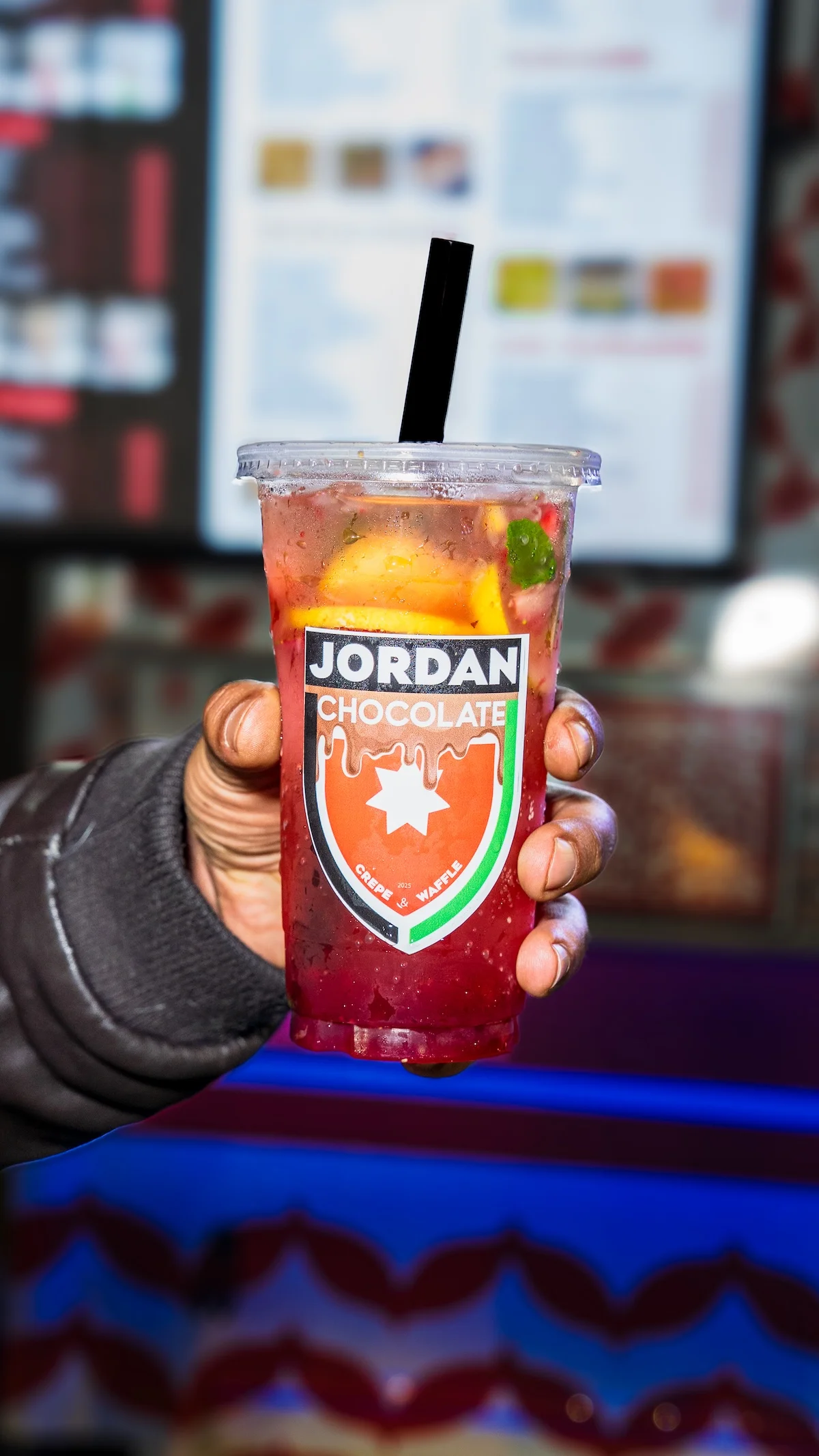 A refreshing beverage in a clear cup, featuring slices of lemon and mint. The drink has a vibrant red color and is labeled with a logo.