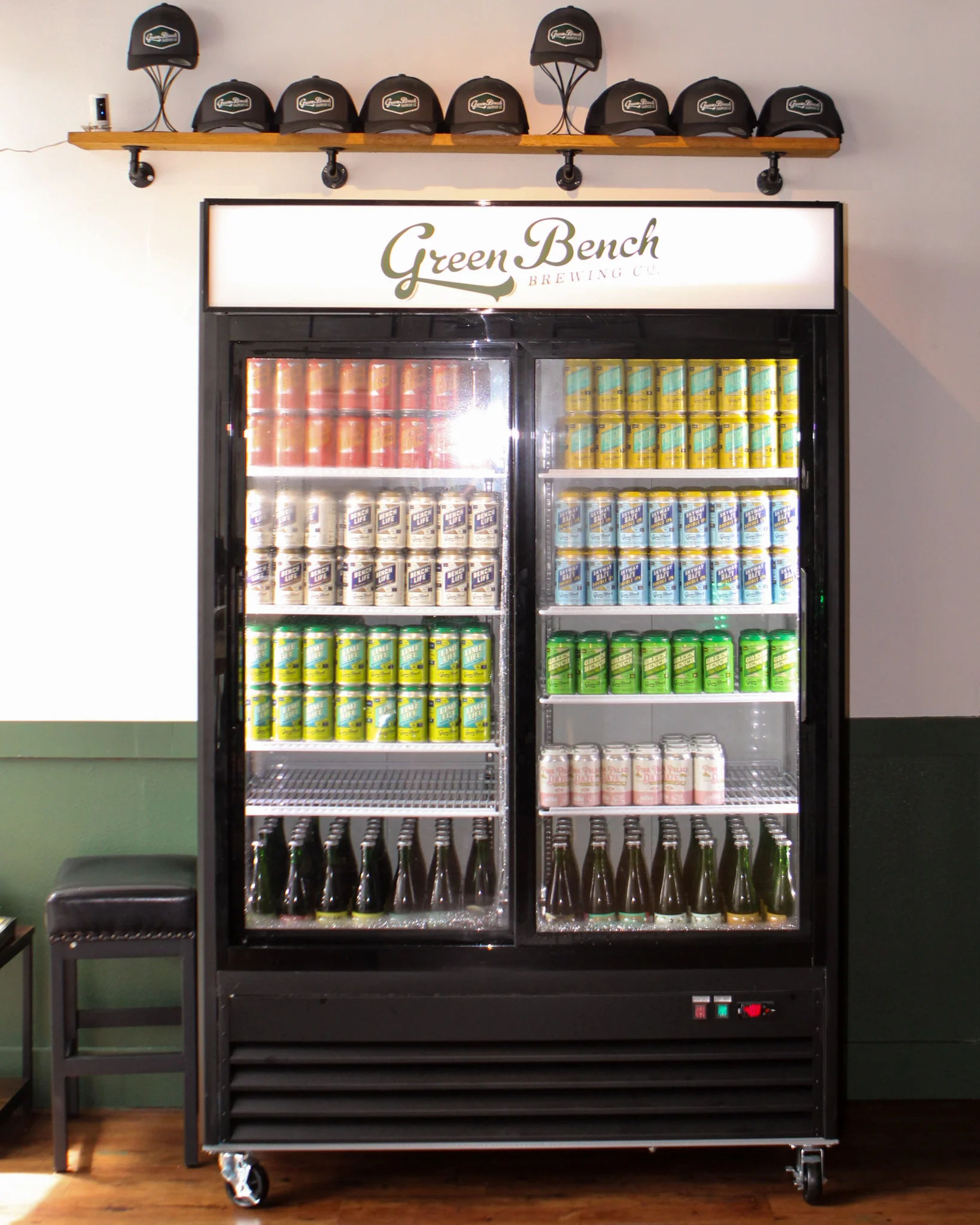 A refrigerator displaying various drinks and cans at a brewery with hats on a shelf above.