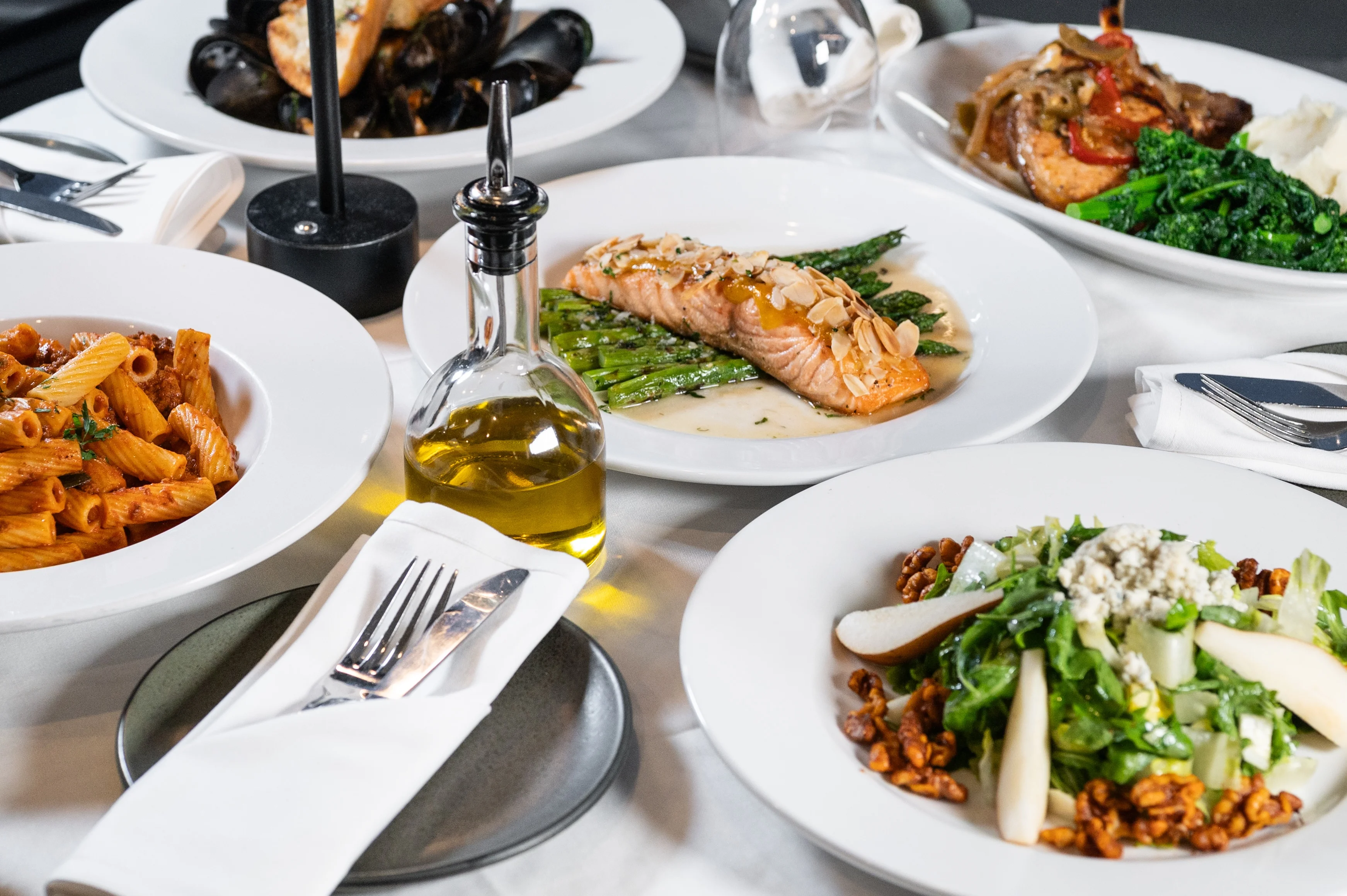 A beautifully arranged table at toscana on main featuring salmon, pasta, salad, and vegetables, with olive oil and cutlery ready for dining.