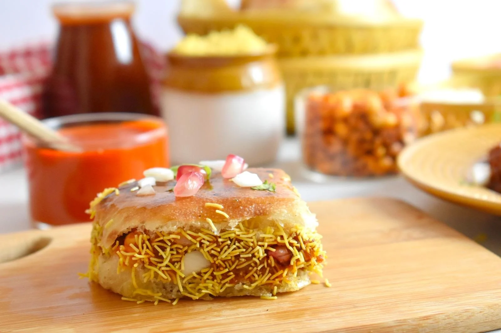A close-up of a savory snack with a crispy layer, filled with tangy ingredients and garnished with onion and pomegranate seeds, served on a wooden board.
