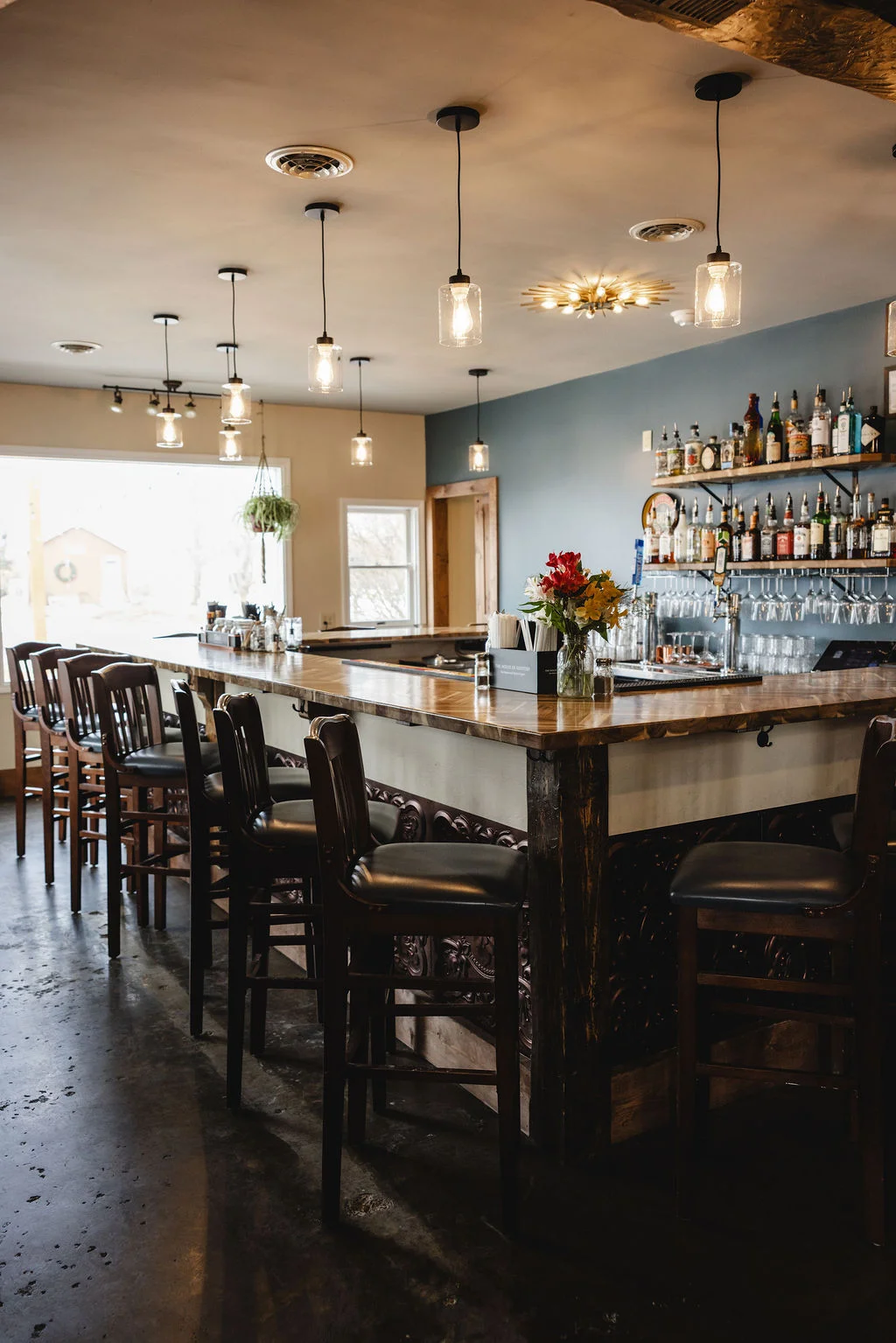 A stylish bar area with wooden stools, decorative lights hanging from the ceiling, and a well-stocked shelf of bottles in the background.