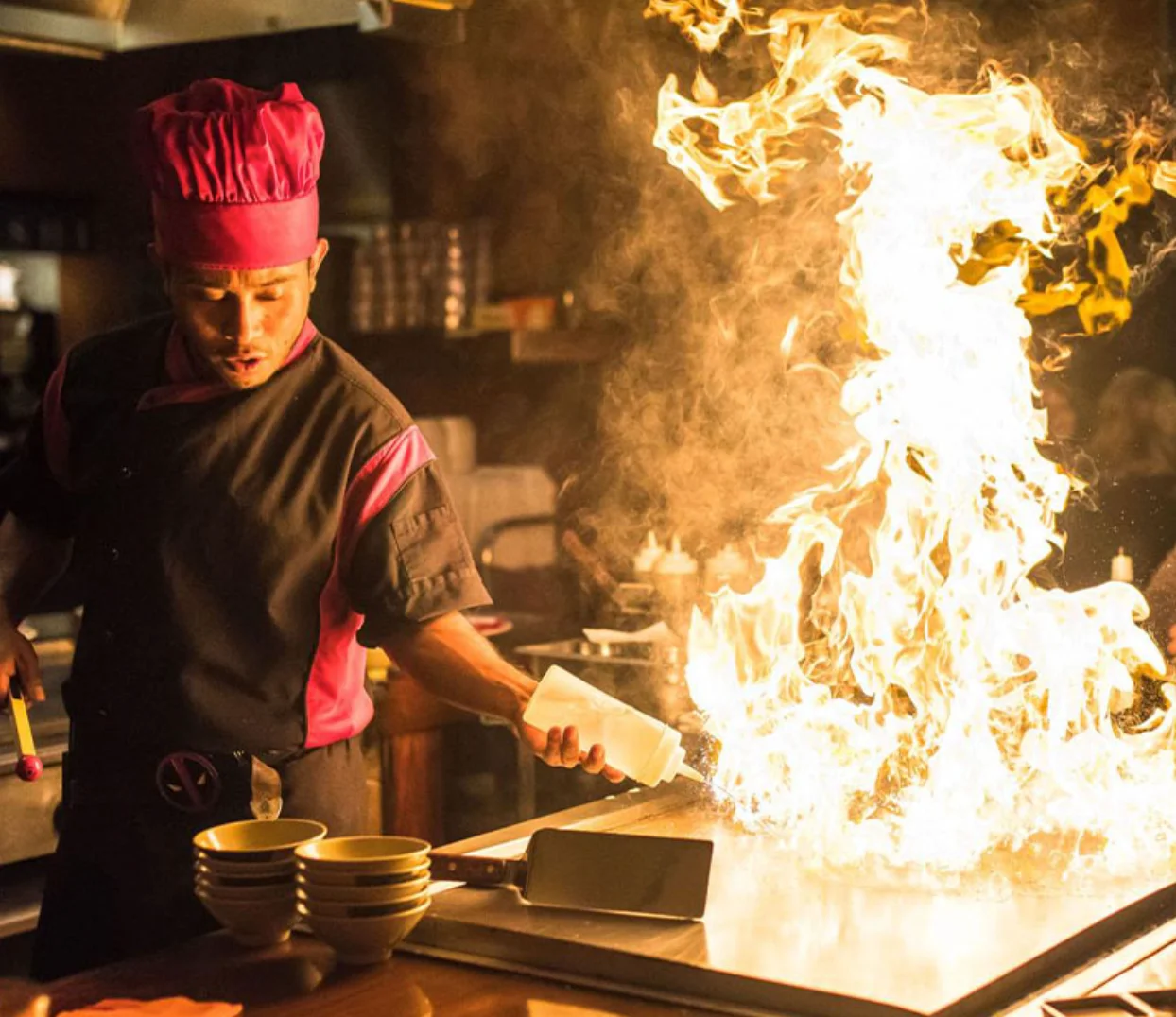 A chef in a pink hat skillfully igniting flames while cooking at a teppanyaki grill. The intense fire creates a dramatic atmosphere in the kitchen.