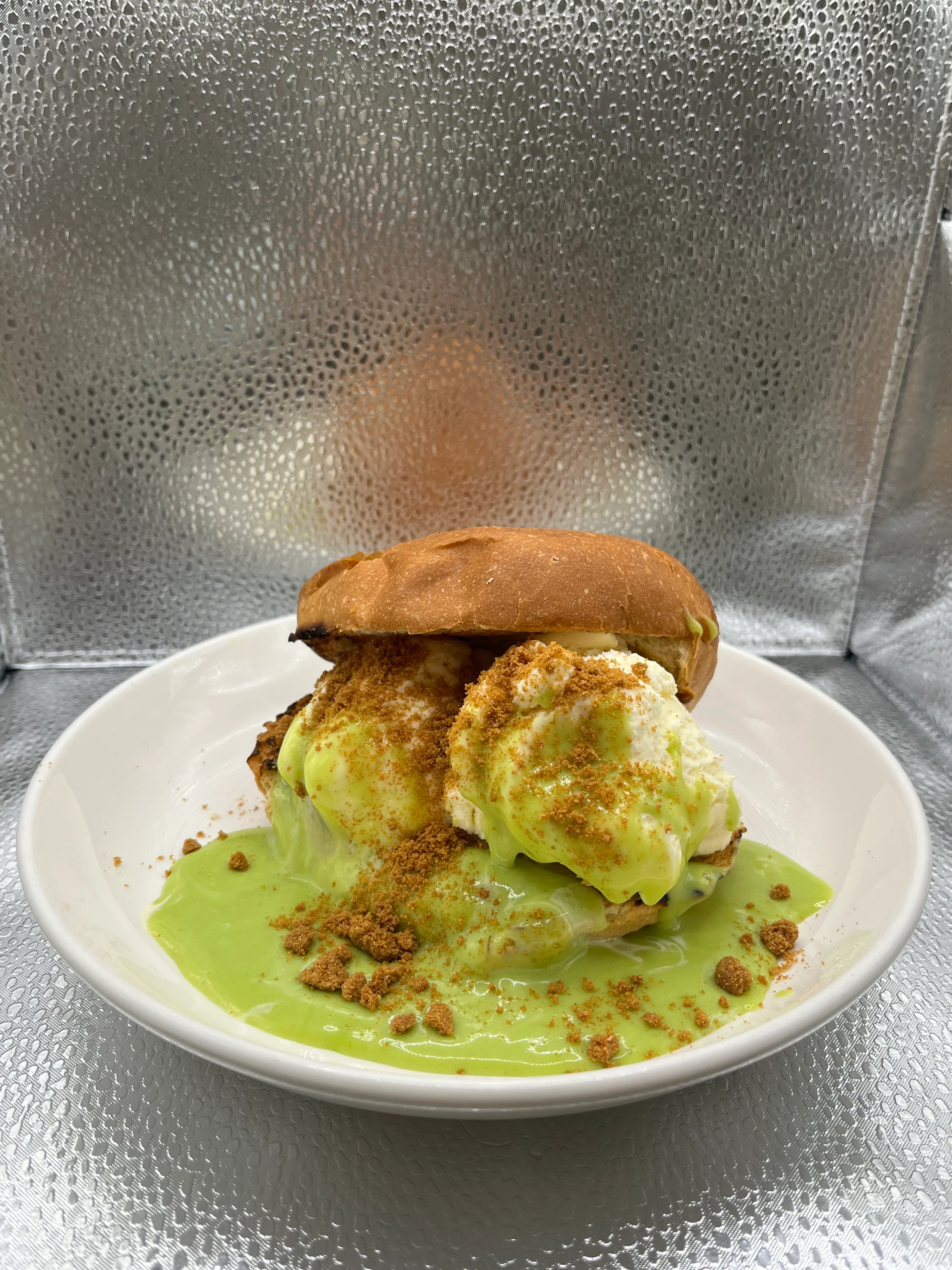 A dessert sandwich made with a soft bun, filled with a creamy filling and topped with crumbled cookies, served on a white plate.