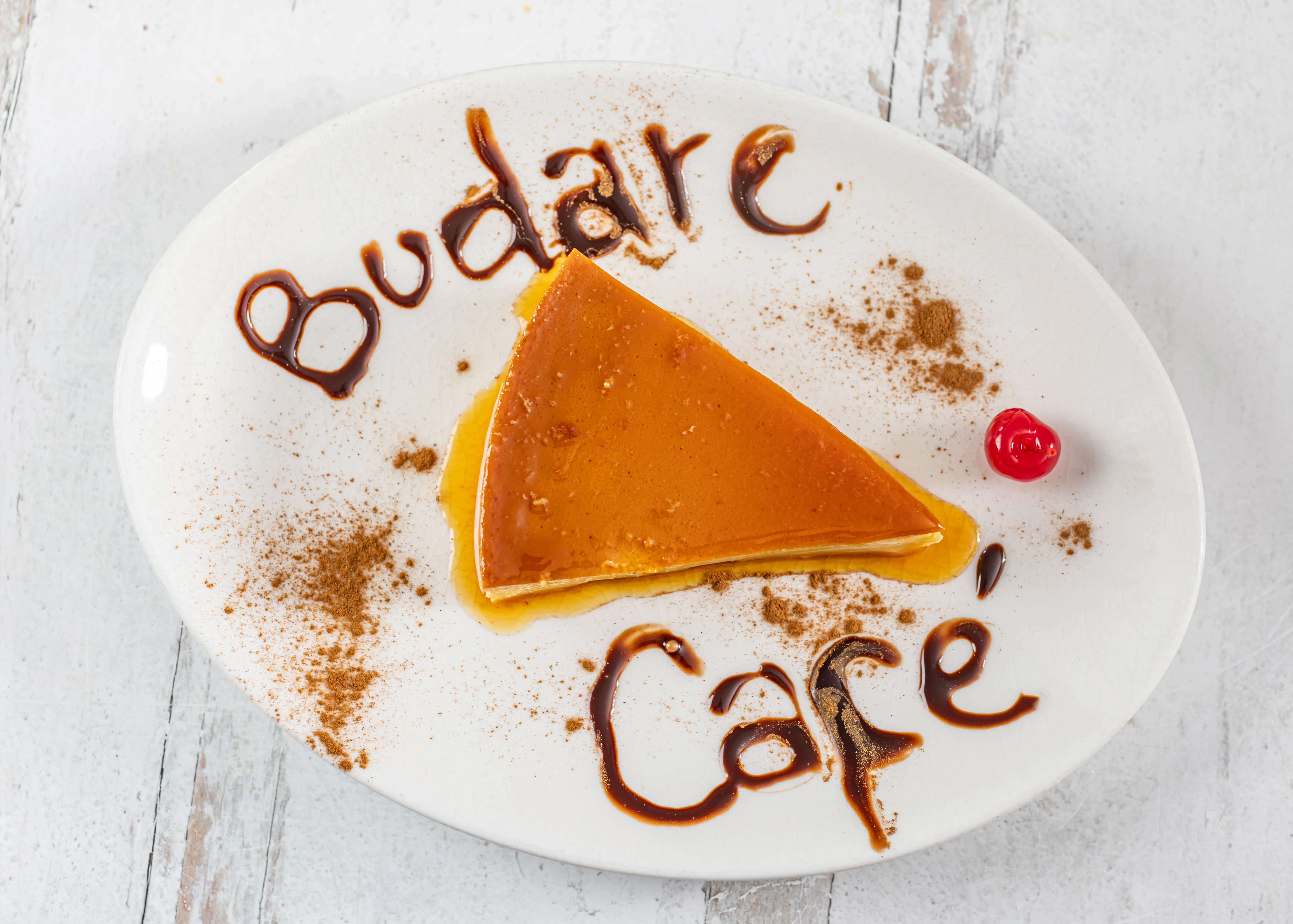 A slice of creamy Venezuelan quesillo (flan), beautifully presented with "Budare Cafe" written in chocolate sauce on the plate.