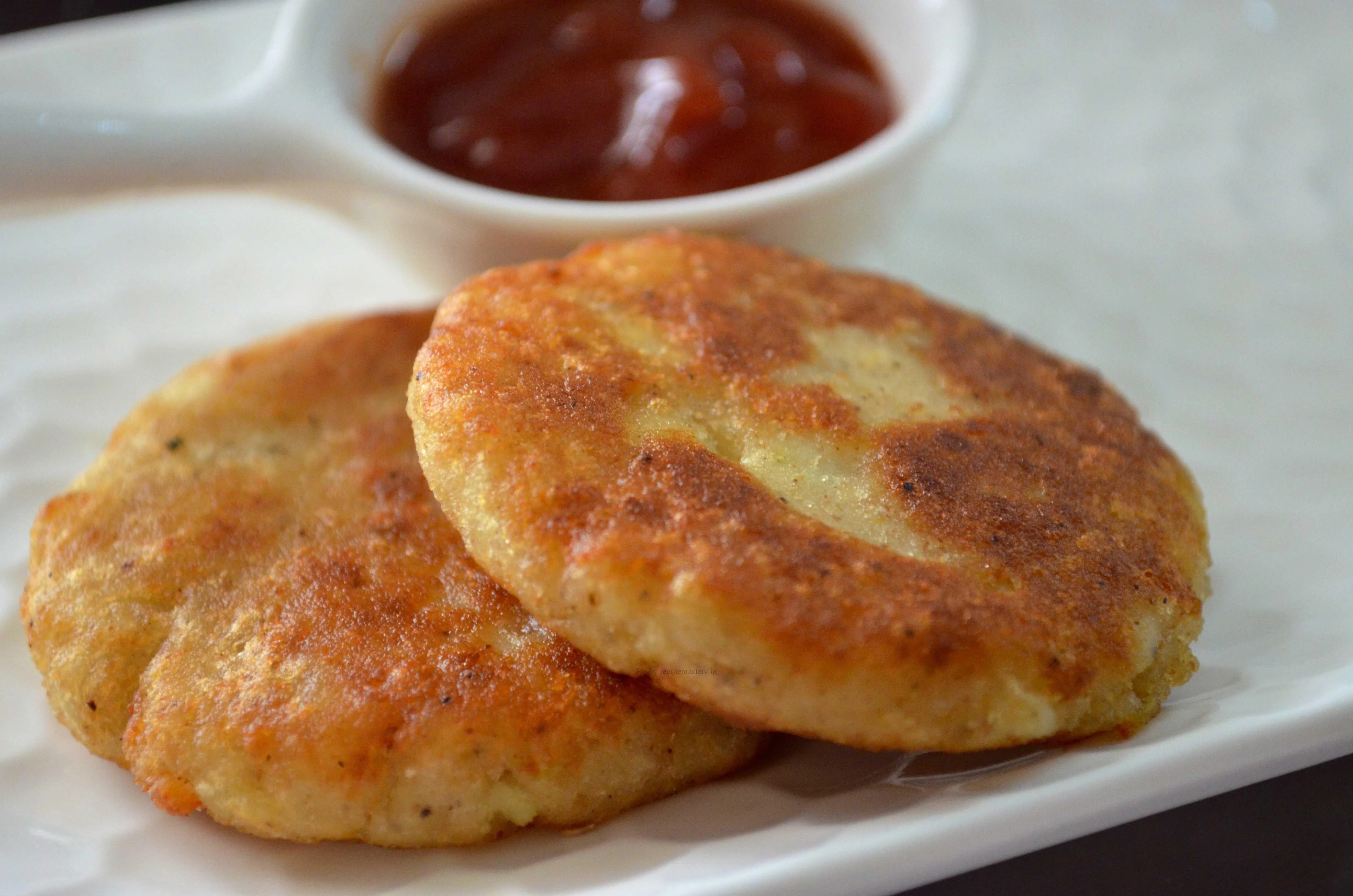 Two golden-brown potato patties served with a small bowl of ketchup on a white plate.