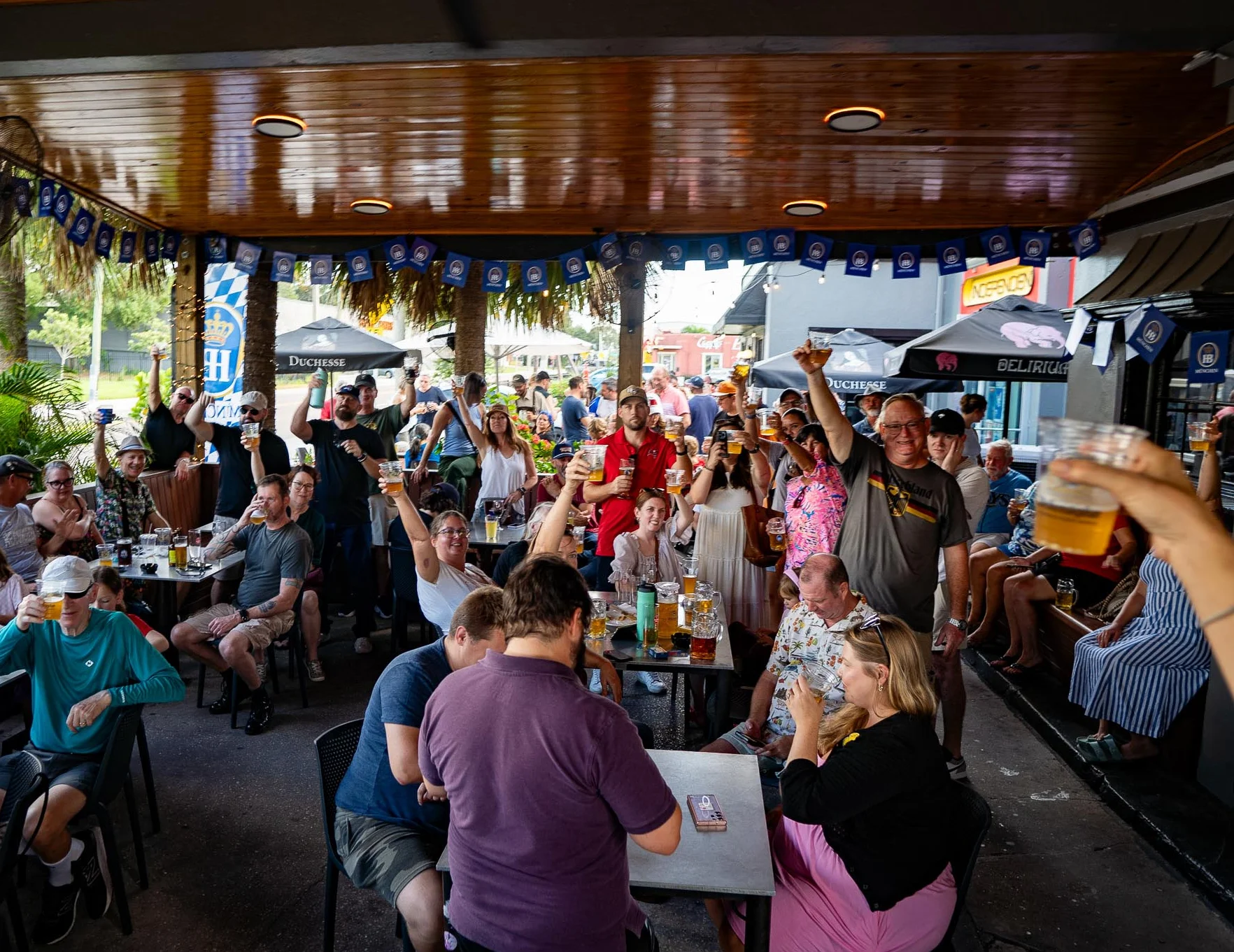 Lively outdoor bar scene with patrons enjoying drinks and raising glasses in celebration.
