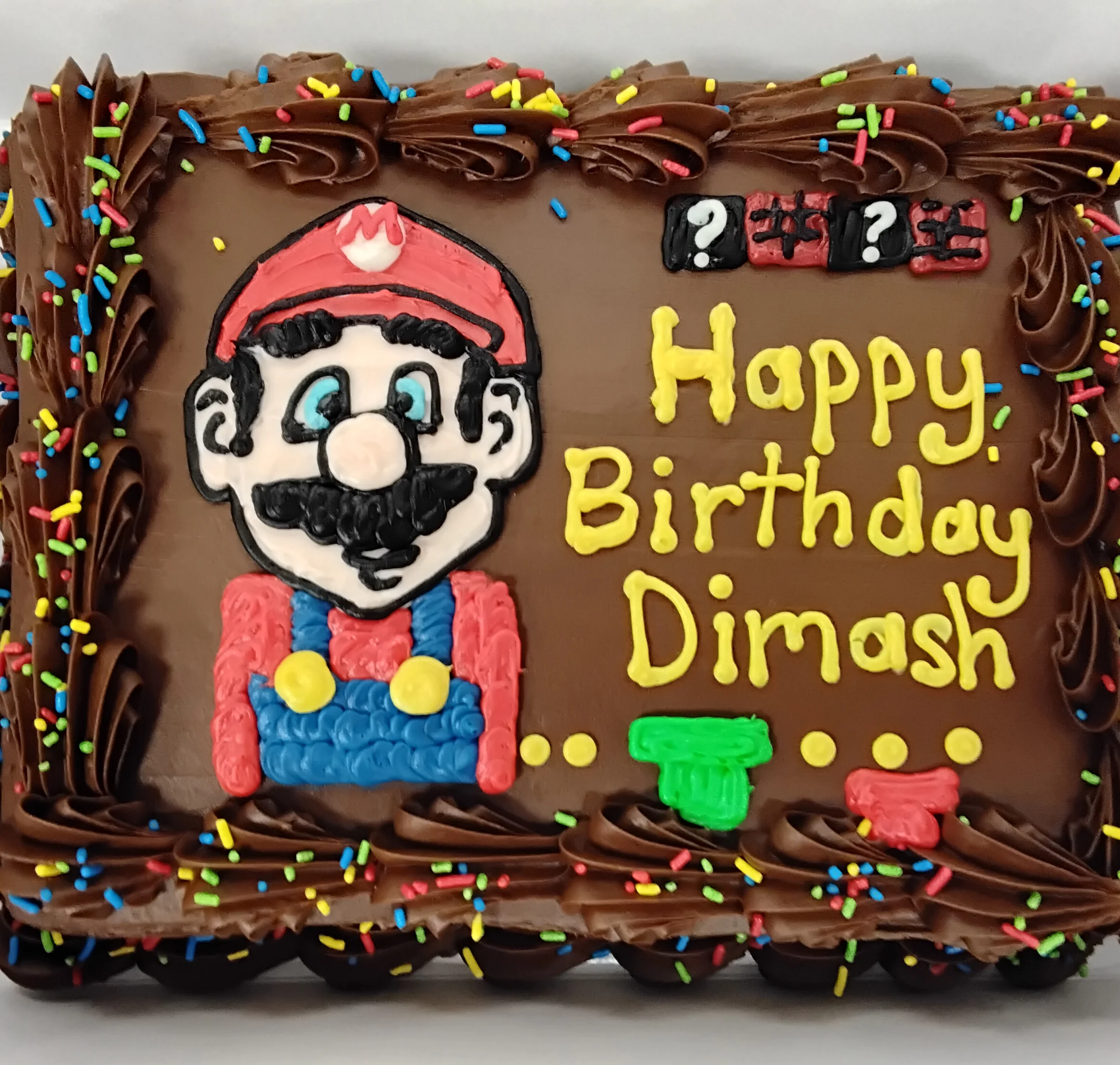 Sheet cake decorated with chocolate and sprinkles with Mario drawn in icing that says "Happy Birthday Dimash"