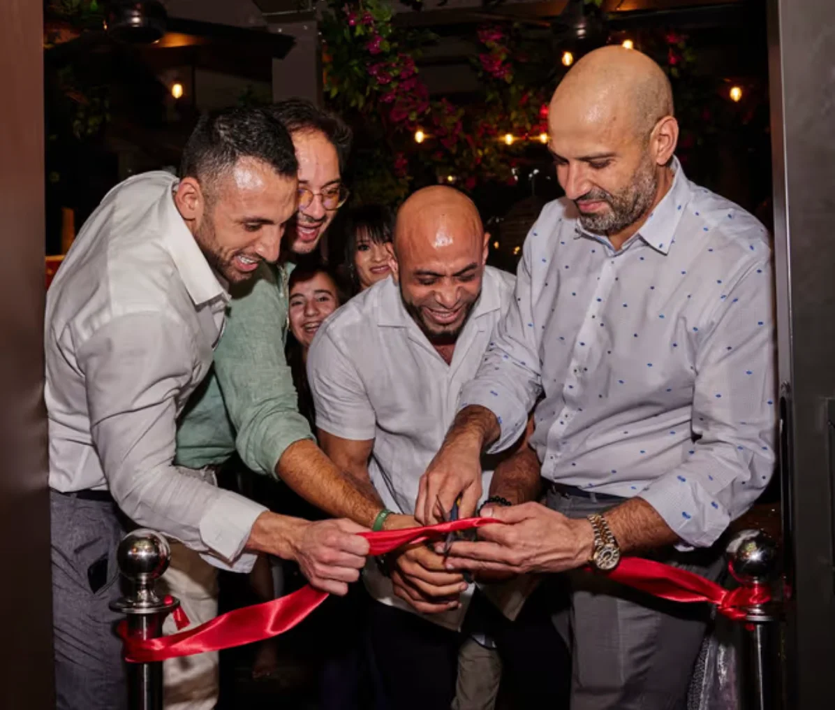 Four men smiling while cutting a red ribbon with a pair of scissors. Two women smiling and peeking through from the dimly lit background behind the men.
