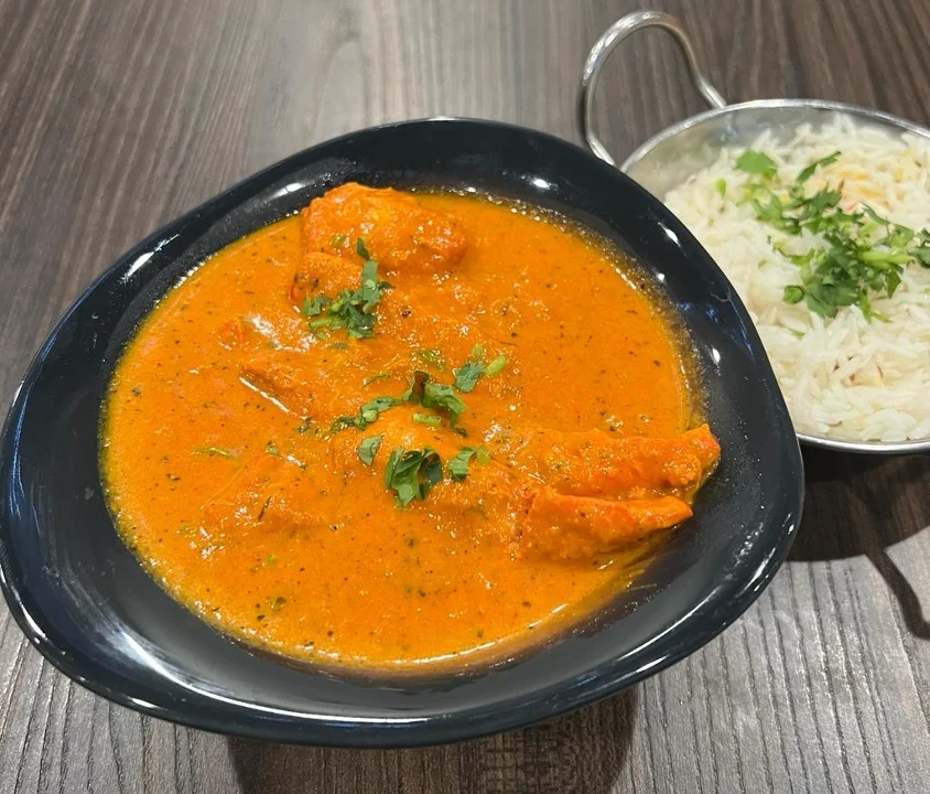 A bowl of curry featuring pieces of meat in a rich, orange sauce, garnished with fresh cilantro. A side of white rice is served in a separate dish.