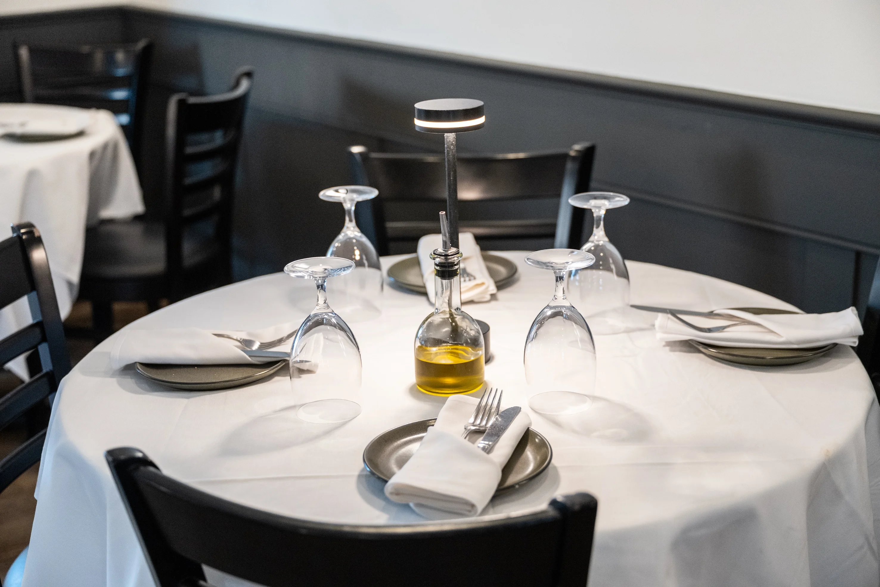 Elegant dining table set with wine glasses, plates, cutlery, and an olive oil bottle under soft lighting, creating a cozy atmosphere.