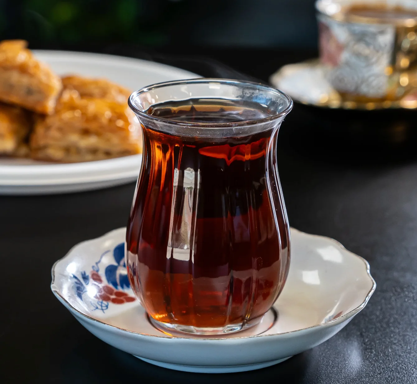 A glass of steaming tea placed on a decorative saucer, with a plate of sweet pastries in the background.