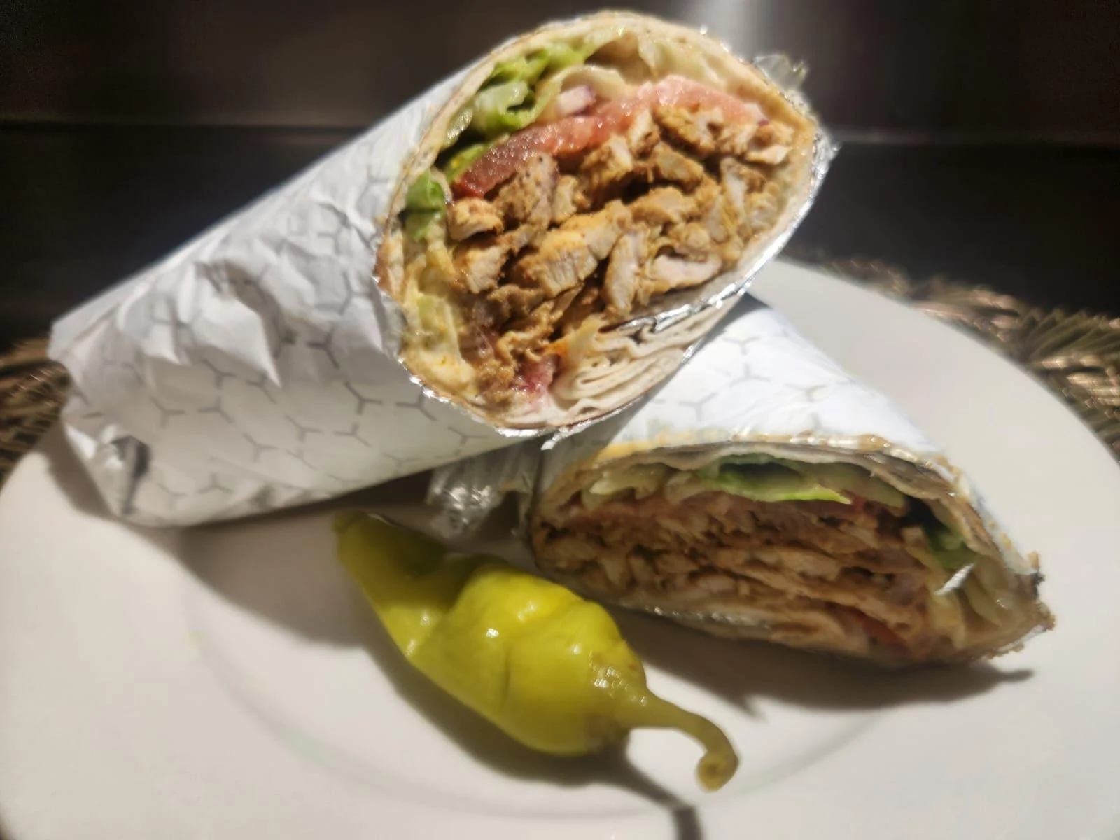 Two wrapped burritos on a plate, showing shredded meat, lettuce, and tomatoes inside. A small chili pepper is placed beside them.