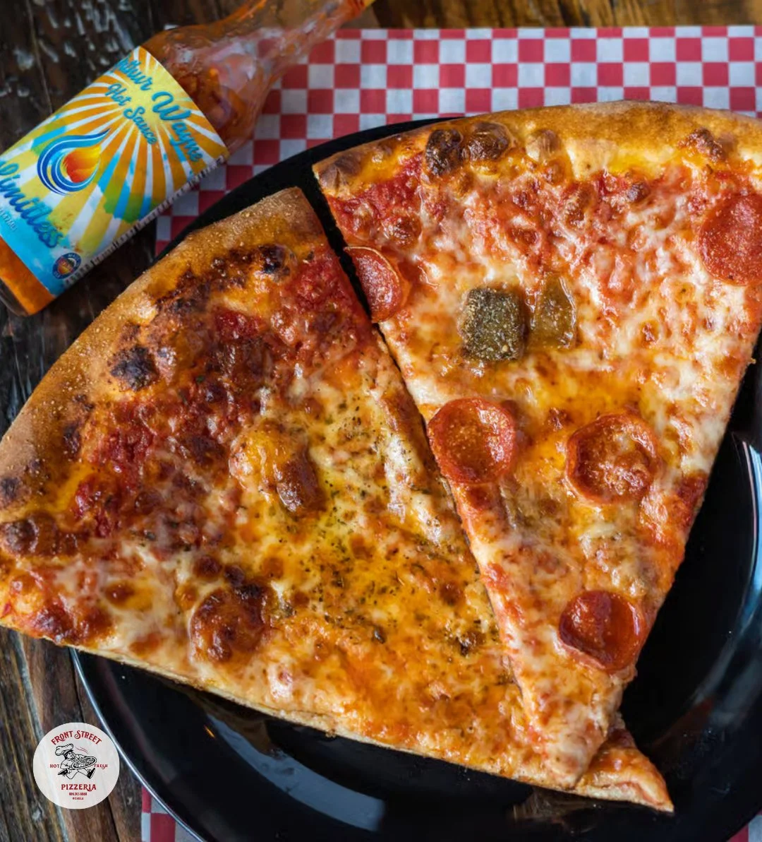 Two slices of pepperoni pizza on a black plate with a bottle of hot sauce in the background.