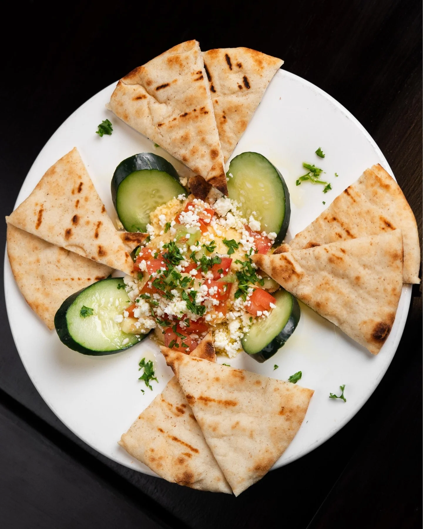 A plate of flatbread wedges surrounding diced tomatoes and cucumbers, topped with herbs and cheese.