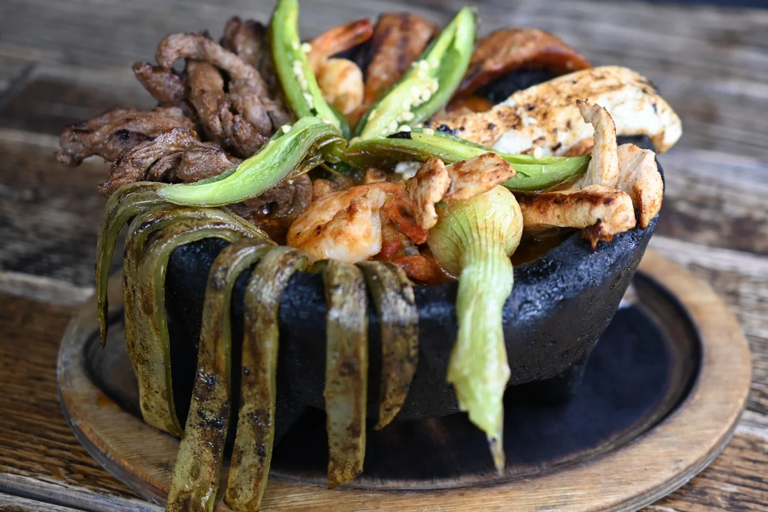 A sizzling dish served in a black stone bowl, featuring a mix of grilled meats and vegetables such as green peppers and onions.