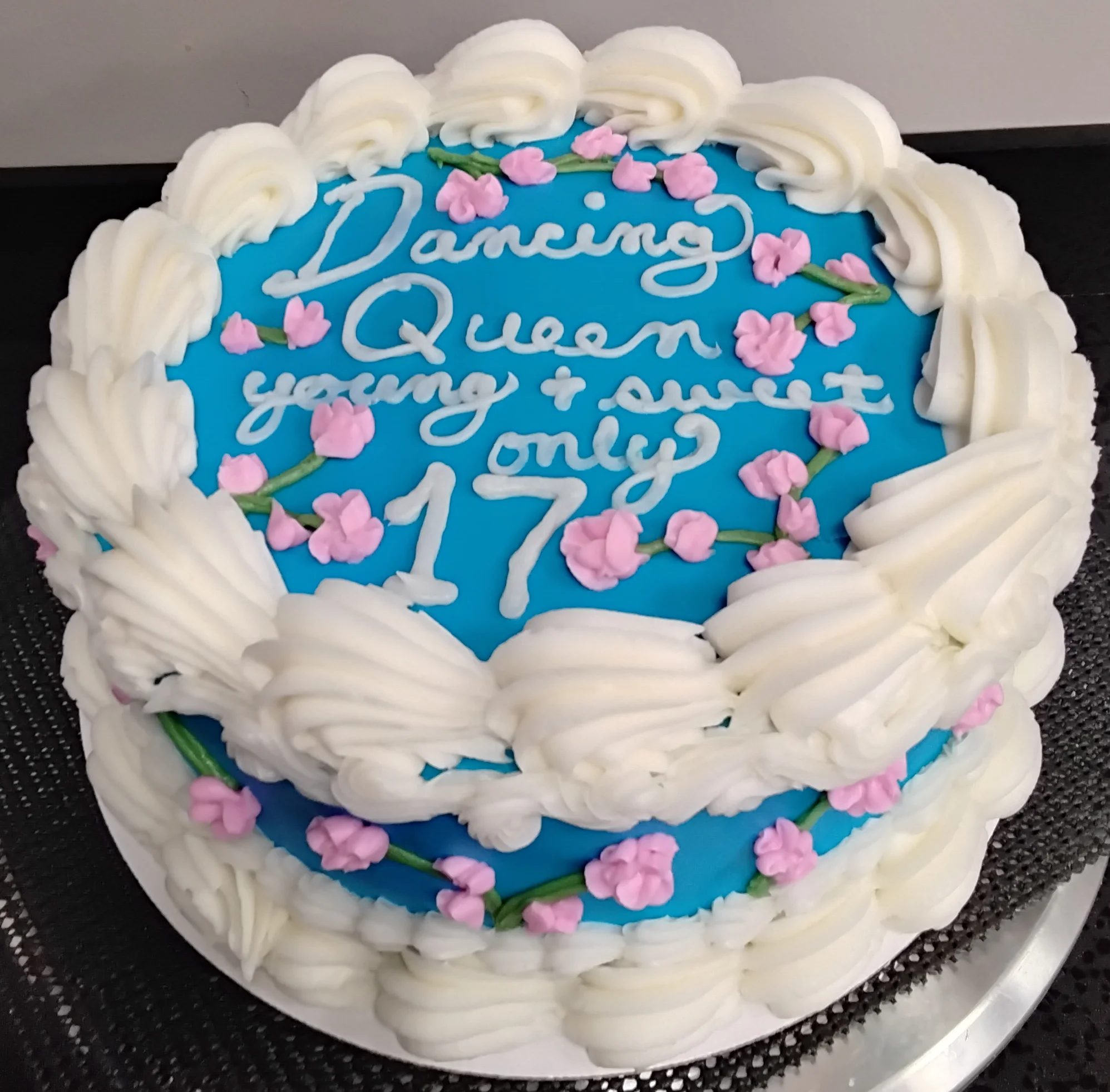 Round cake with blue/teal icing and white borders. Decorated with green stems and small pink flowers and the words "Dancing Queen, young & sweet only 17"