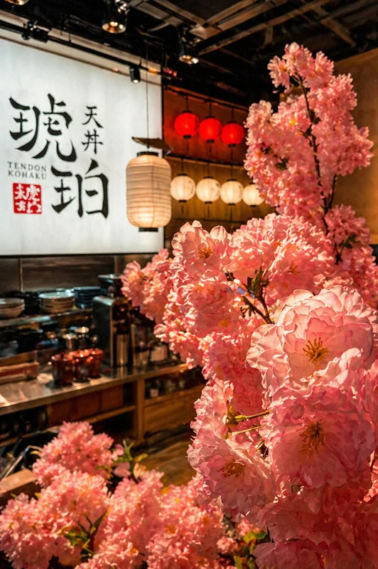 A vibrant interior view of Tendon Kohaku framed by a large branch of pink cherry blossoms in the immediate foreground. The background opens up to a kitchen counter, hanging traditional white and red lanterns, and a massive backlit wall panel featuring the restaurant’s bold black Japanese calligraphy and red stamp logo.