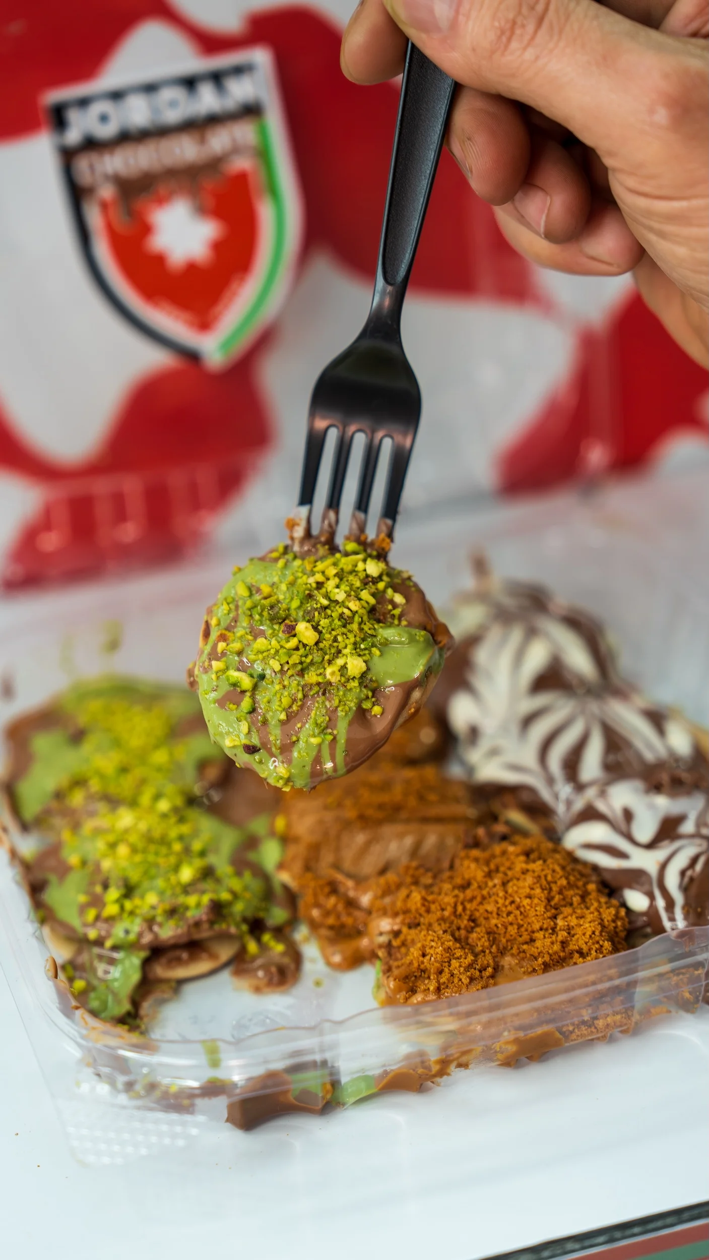 A fork holding a bite-sized treat topped with green sauce and crushed nuts, with various desserts in the background.