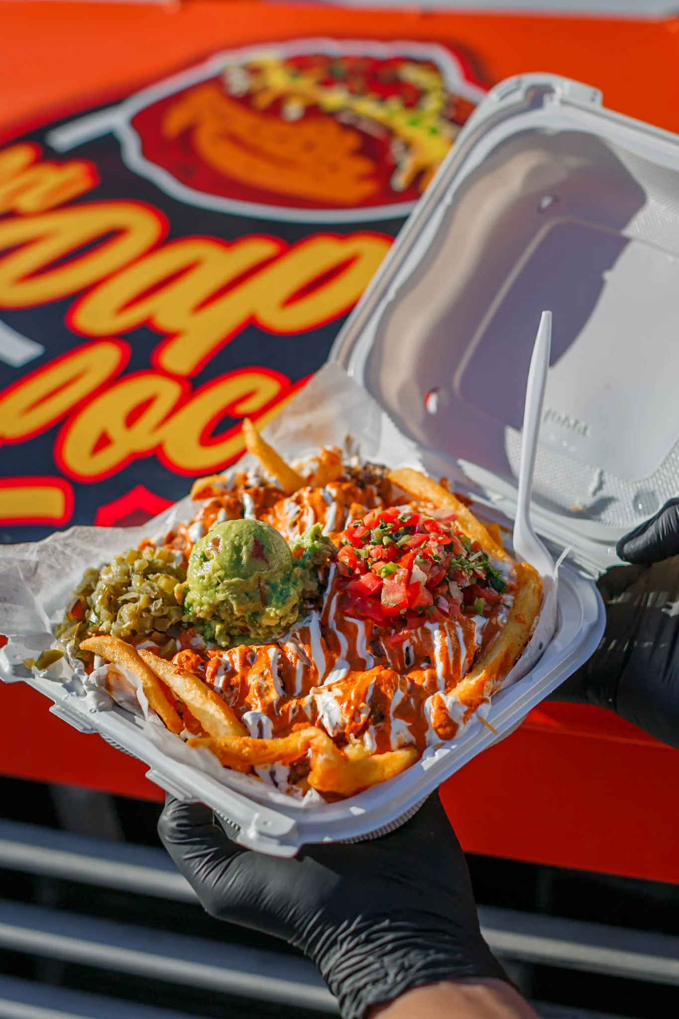 A container of loaded fries topped with meat, guacamole, fresh salsa, and a drizzle of sauce, accompanied by jalapeños.