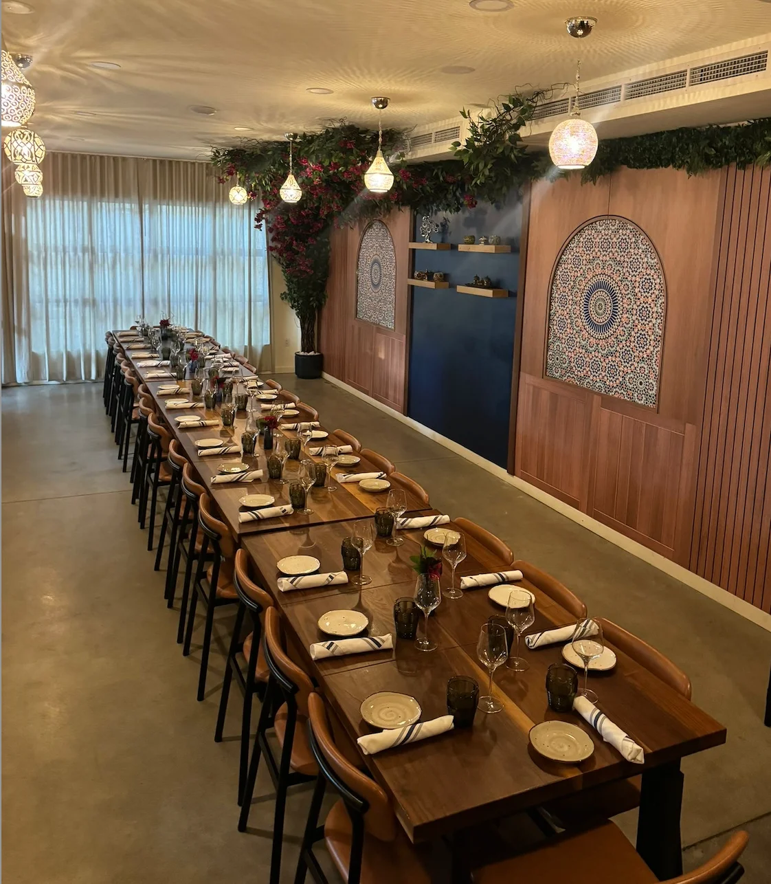 Long dining table set for a formal event with ornate plates, wine glasses, and folded napkins. Elegant lighting, decorative wall with mosaic patterns.
