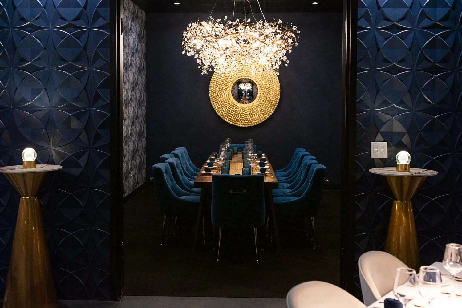 Elegant private dining room with a long wooden table surrounded by plush blue velvet chairs, set with glassware and navy napkins. A striking gold circular mirror and crystal chandelier add a touch of glamour, with dark patterned walls and modern gold accent tables completing the sophisticated ambiance.