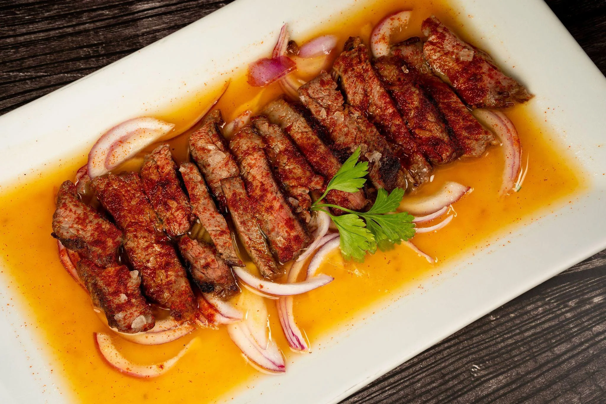Slices of seasoned beef arranged on a white plate, garnished with onions and herbs in a savory sauce.