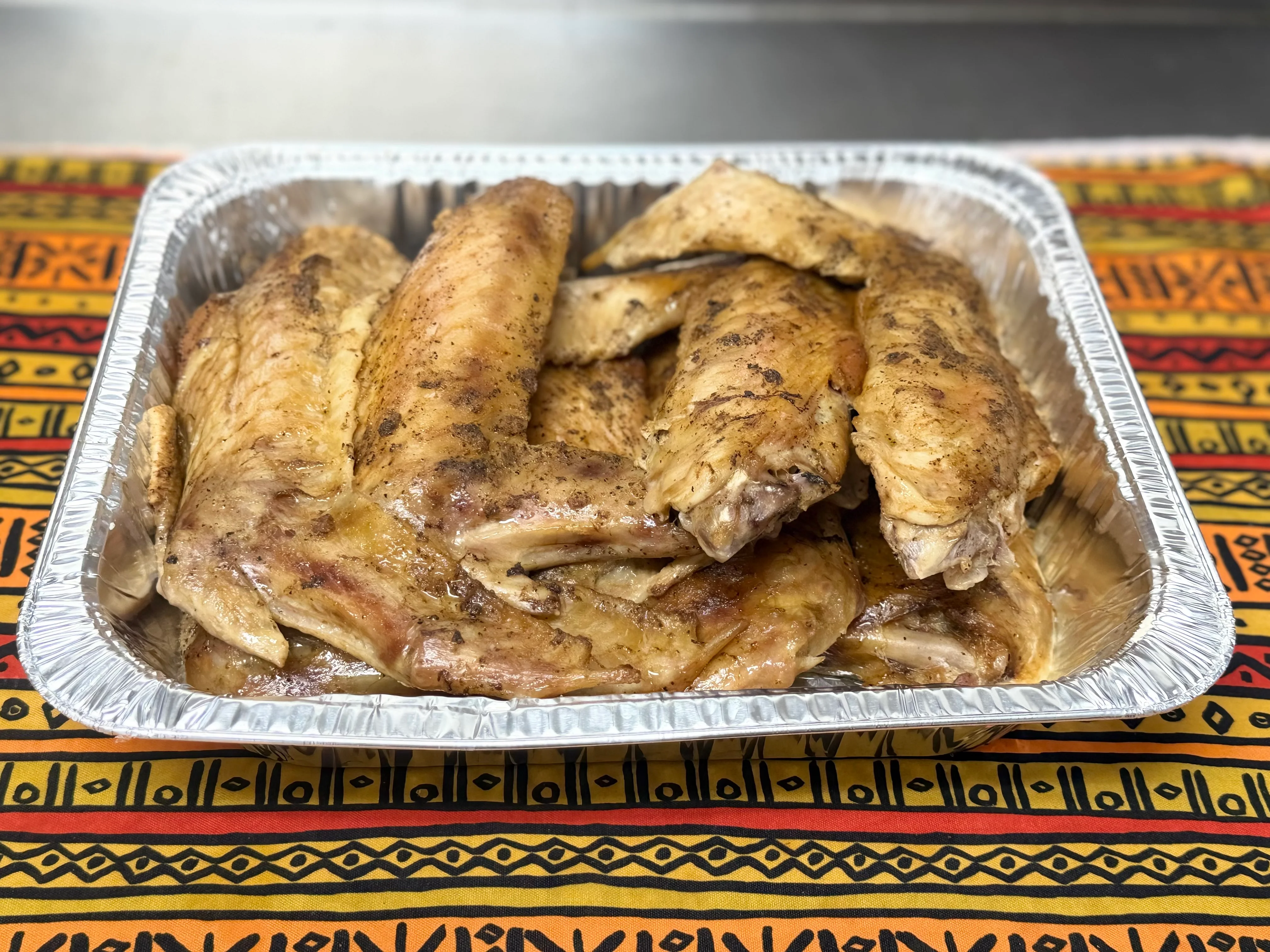 A tray of seasoned, cooked chicken wings stacked in an aluminum container, resting on a vibrant patterned tablecloth.