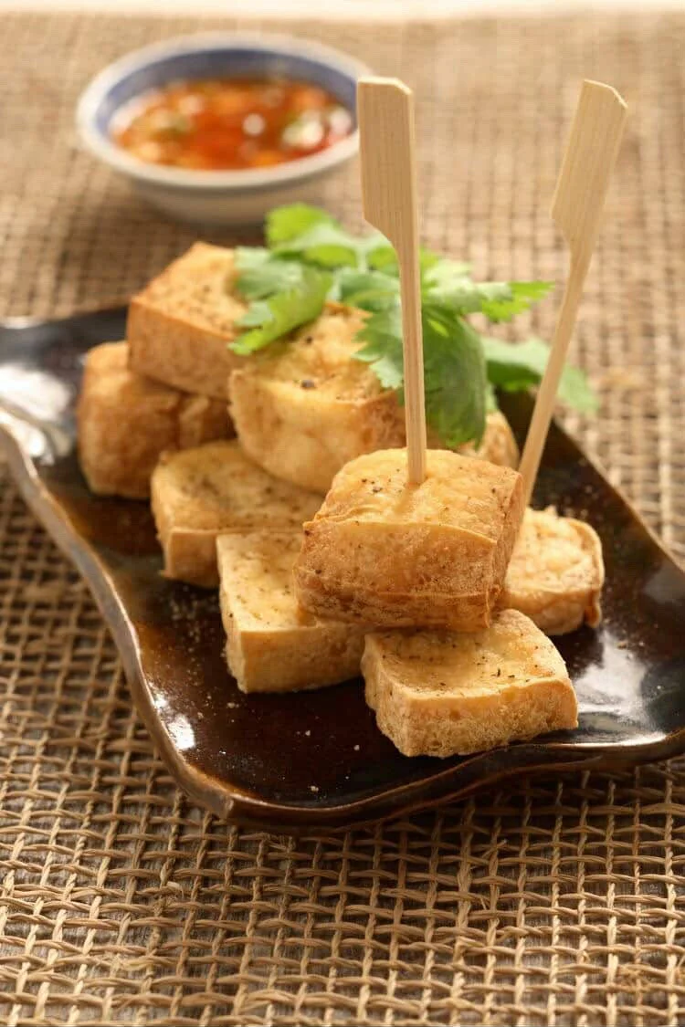 A serving of fried tofu pieces garnished with cilantro and served on a dark plate alongside a dipping sauce.