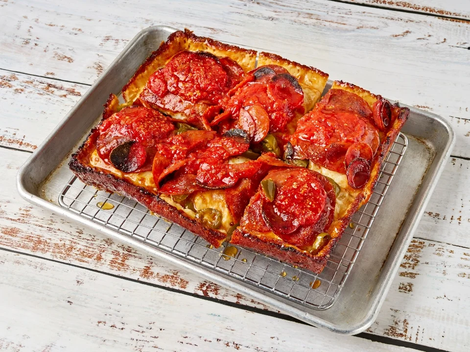 A rectangular metal tray holding a thick crust pizza topped with pepperoni and a rich marinara sauce.
