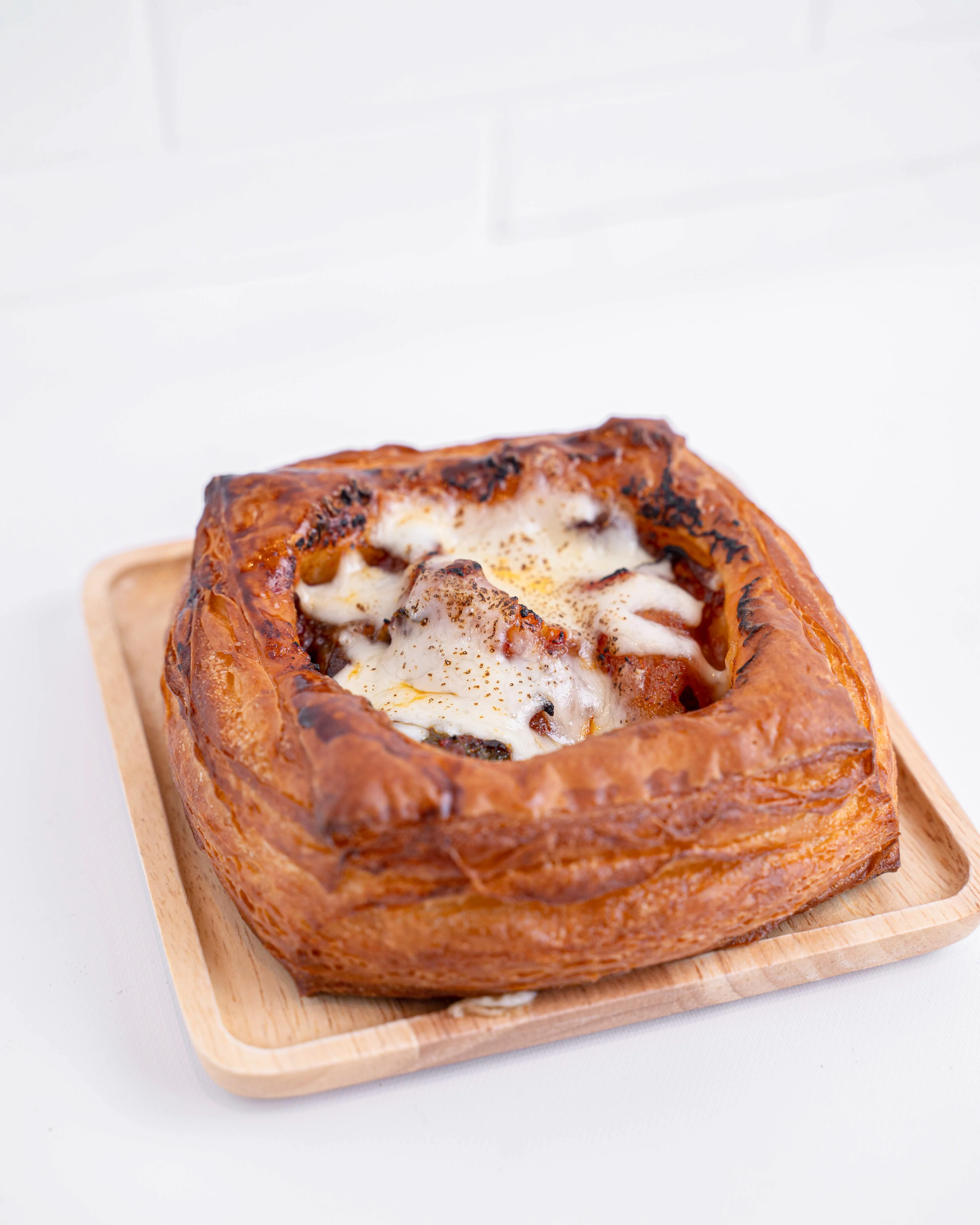 A square pastry topped with melted cheese, presented on a wooden plate against a light backdrop.
