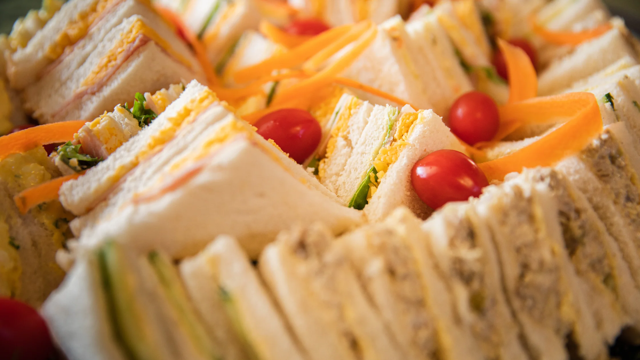 Close-up of assorted finger sandwiches made with white bread, layered with fillings like shredded carrots, sliced cucumber, ham, egg salad, and cheddar cheese, garnished with cherry tomatoes and fresh greens