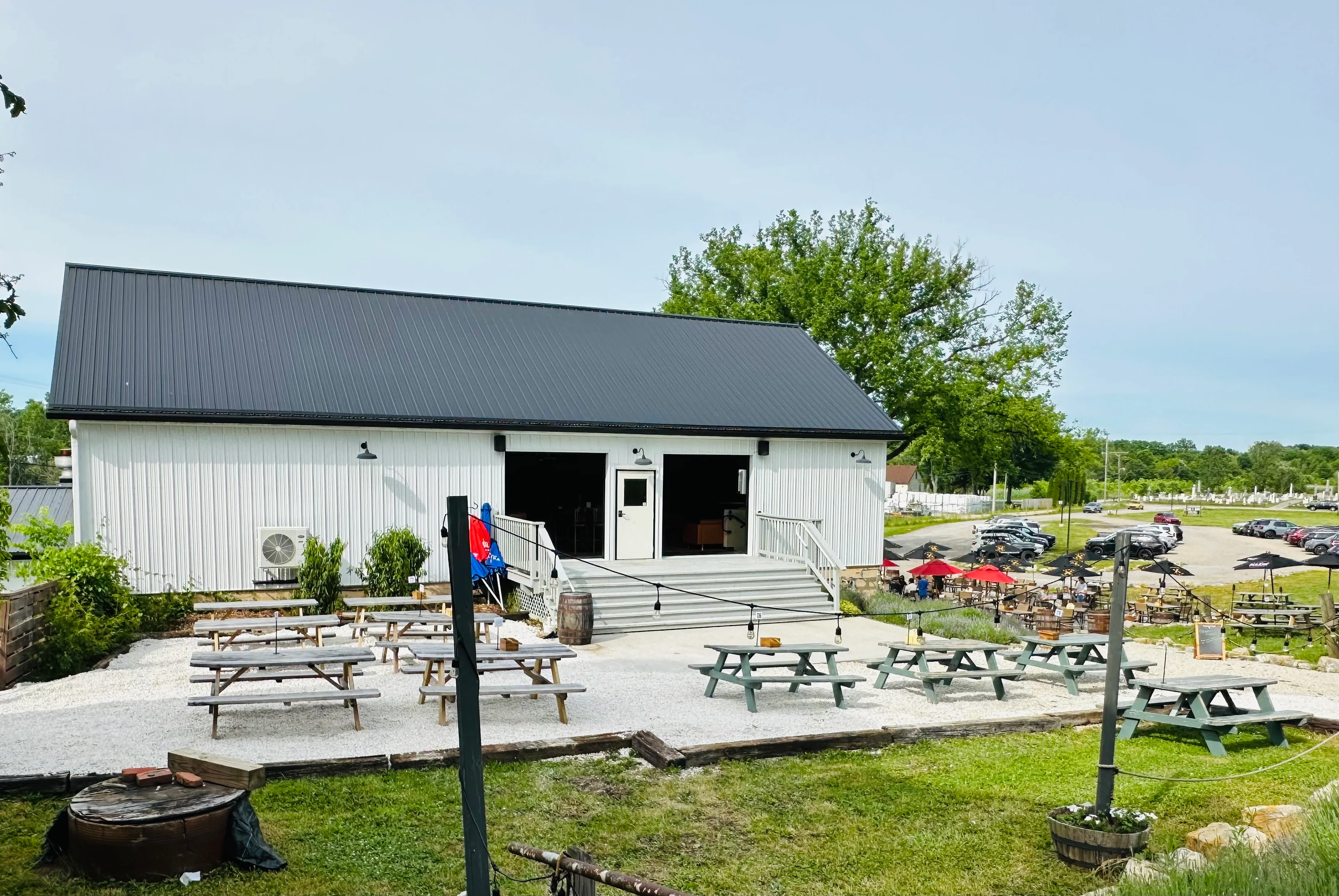 A white building with a black roof and outdoor seating area, featuring picnic tables and a large parking lot.