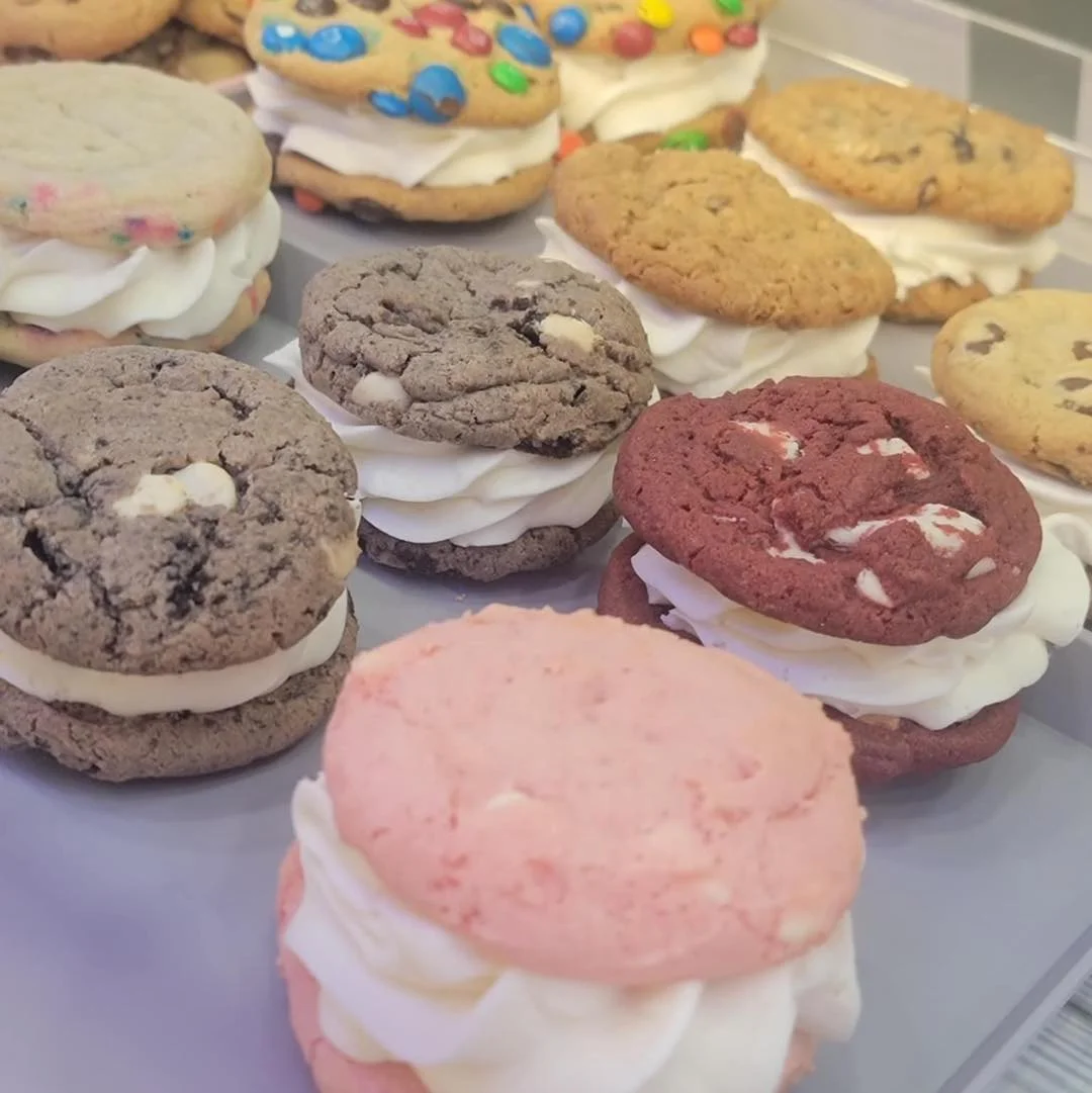 Cookie sandwiches in different flavors, including chocolate, red velvet, and funfetti, filled with white cream frosting.