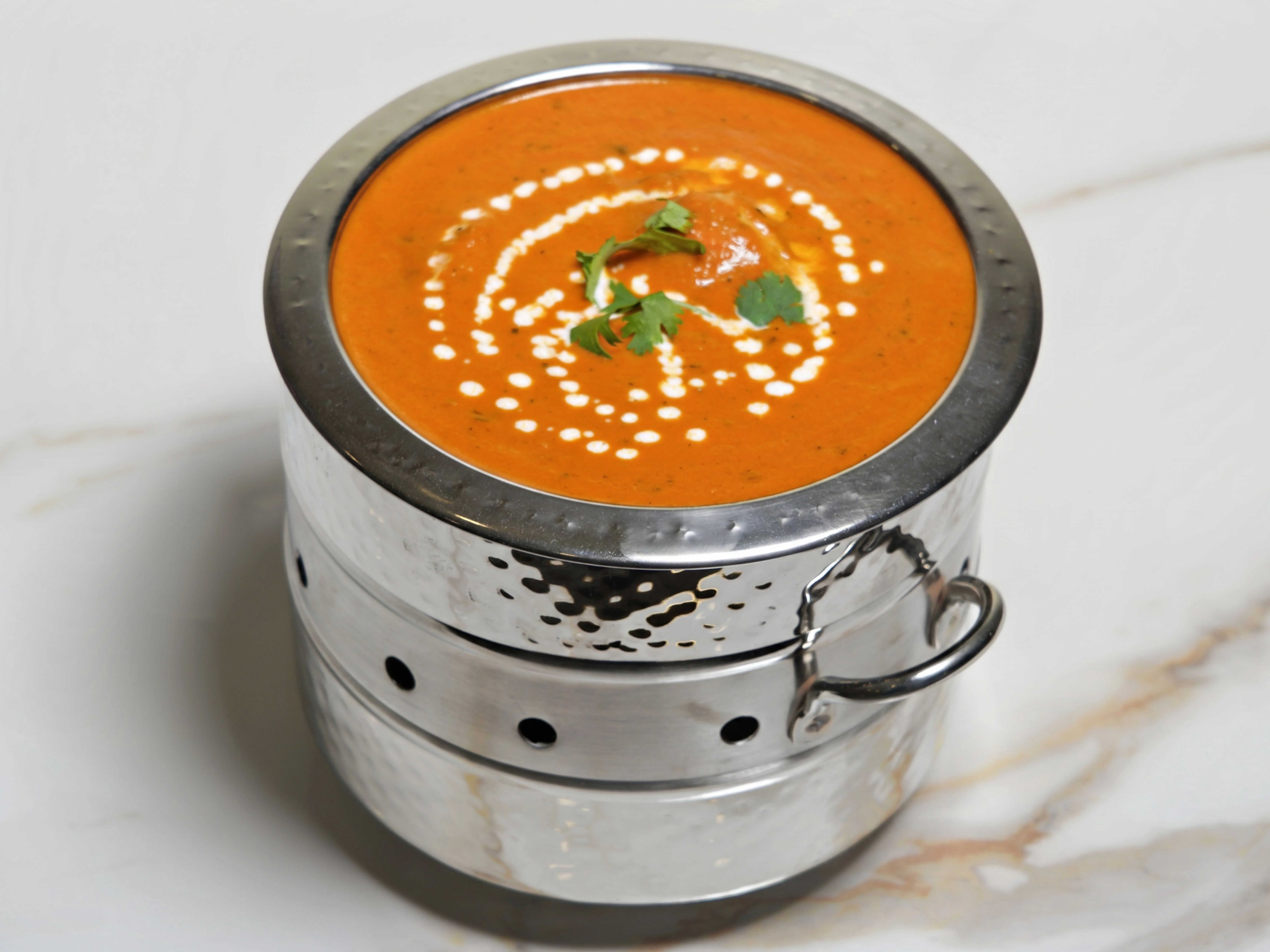 A vibrant orange soup served in a two-tiered silver container, drizzled with cream and garnished with herbs.