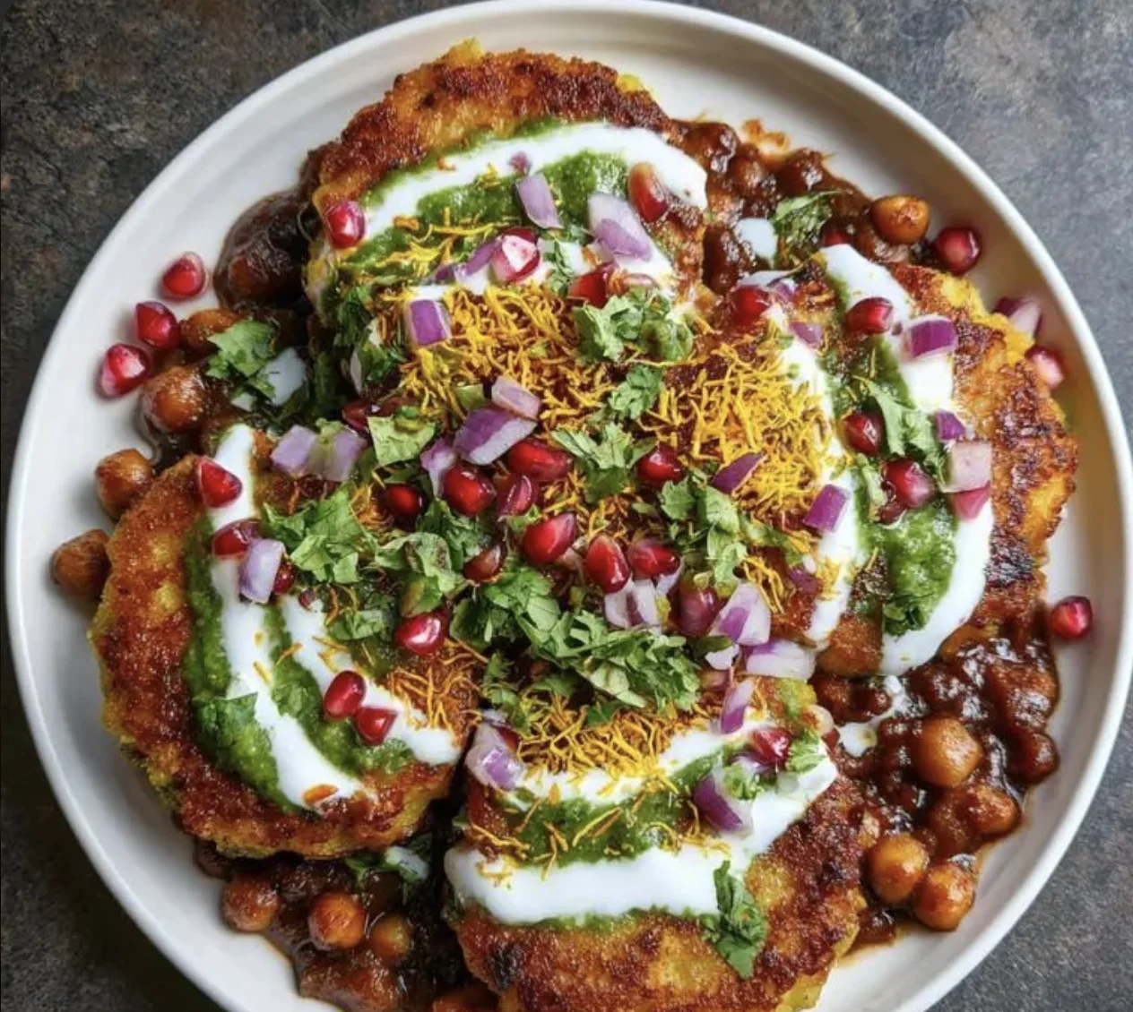 A colorful dish featuring crispy fried patties topped with yogurt, green sauce, and garnished with pomegranate seeds, onions, and fried noodles.