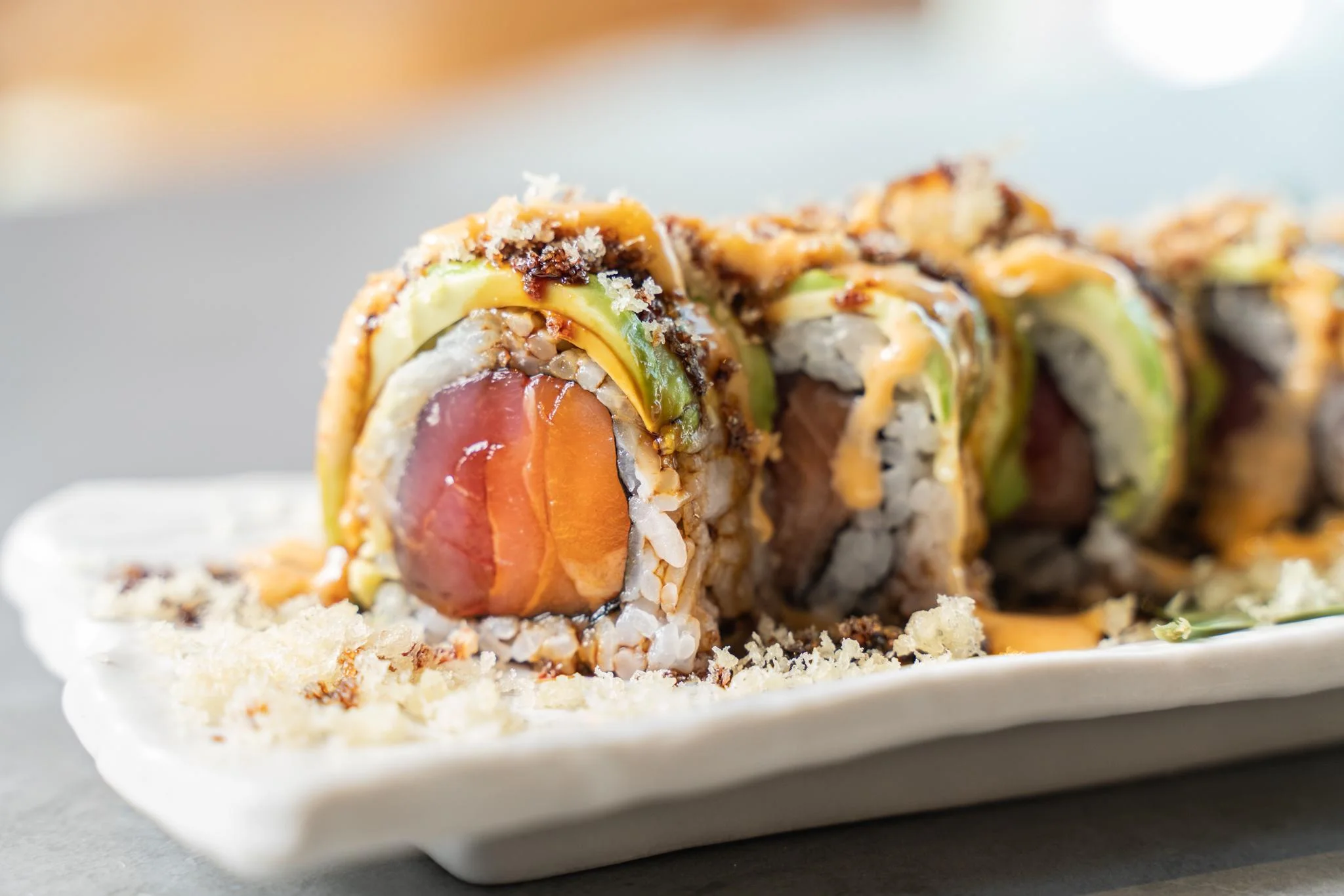 A close-up of a sushi roll topped with crunchy ingredients and drizzled with a savory sauce, highlighting the layers of fish and vegetables.