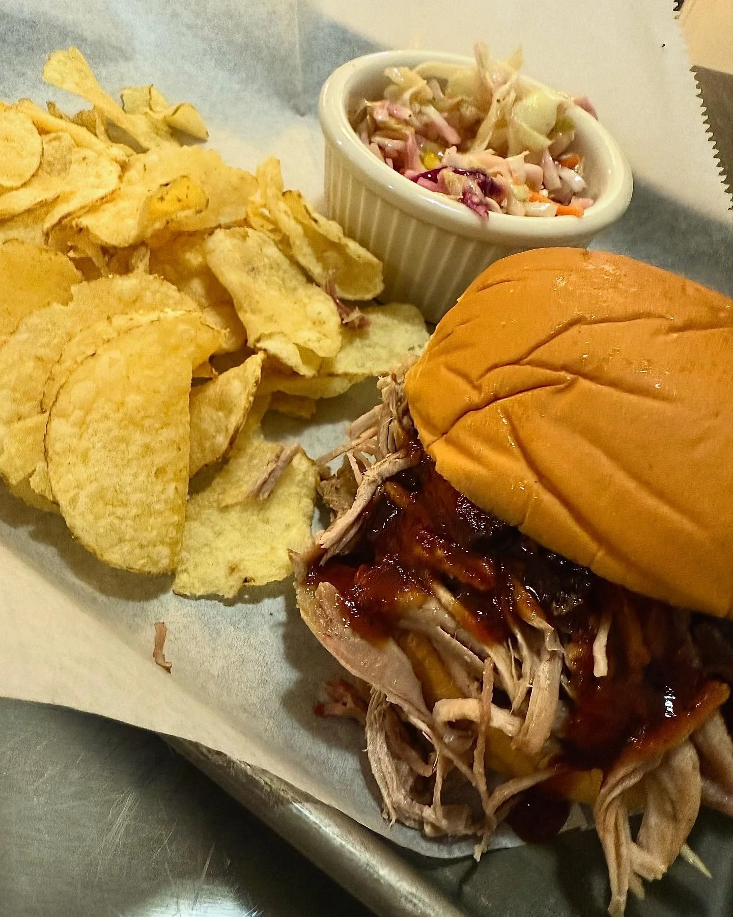 A plate featuring a pulled pork sandwich with barbecue sauce, accompanied by crispy potato chips and a small bowl of coleslaw.