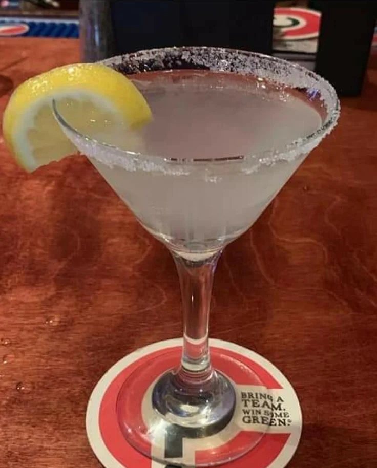 A classic cocktail in a glass with a salted rim, garnished with a lemon wedge, resting on a bar counter.