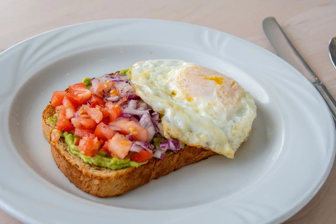 A slice of whole grain toast topped with avocado, diced tomatoes, red onions, and a sunny-side-up egg.