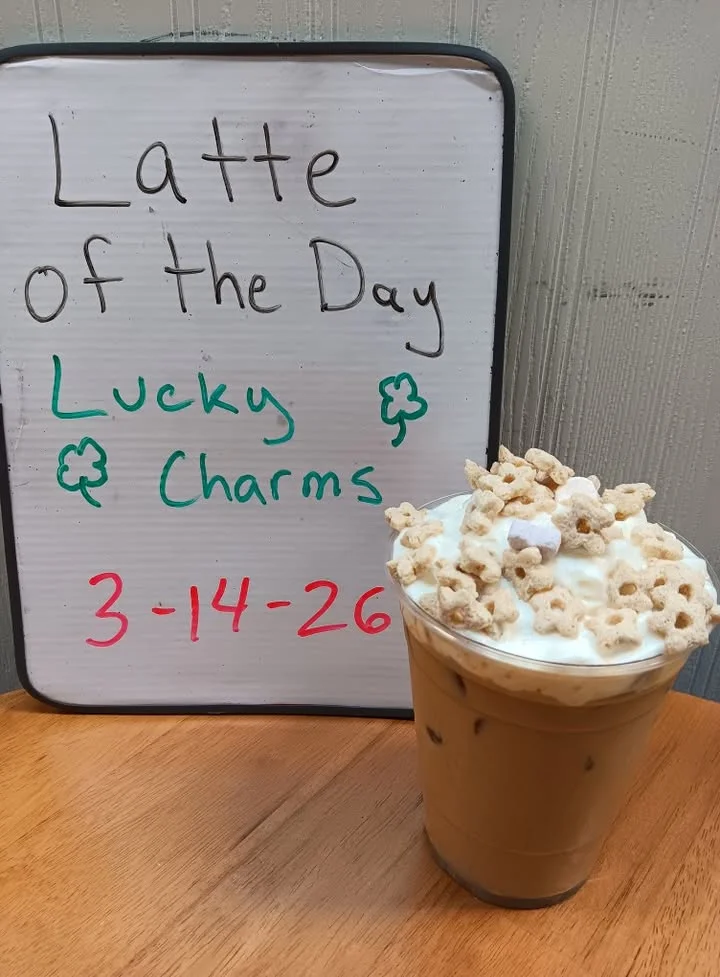 A promotional sign for a latte of the day named 'Lucky Charms,' alongside a tall glass of iced latte topped with whipped cream and cereal.