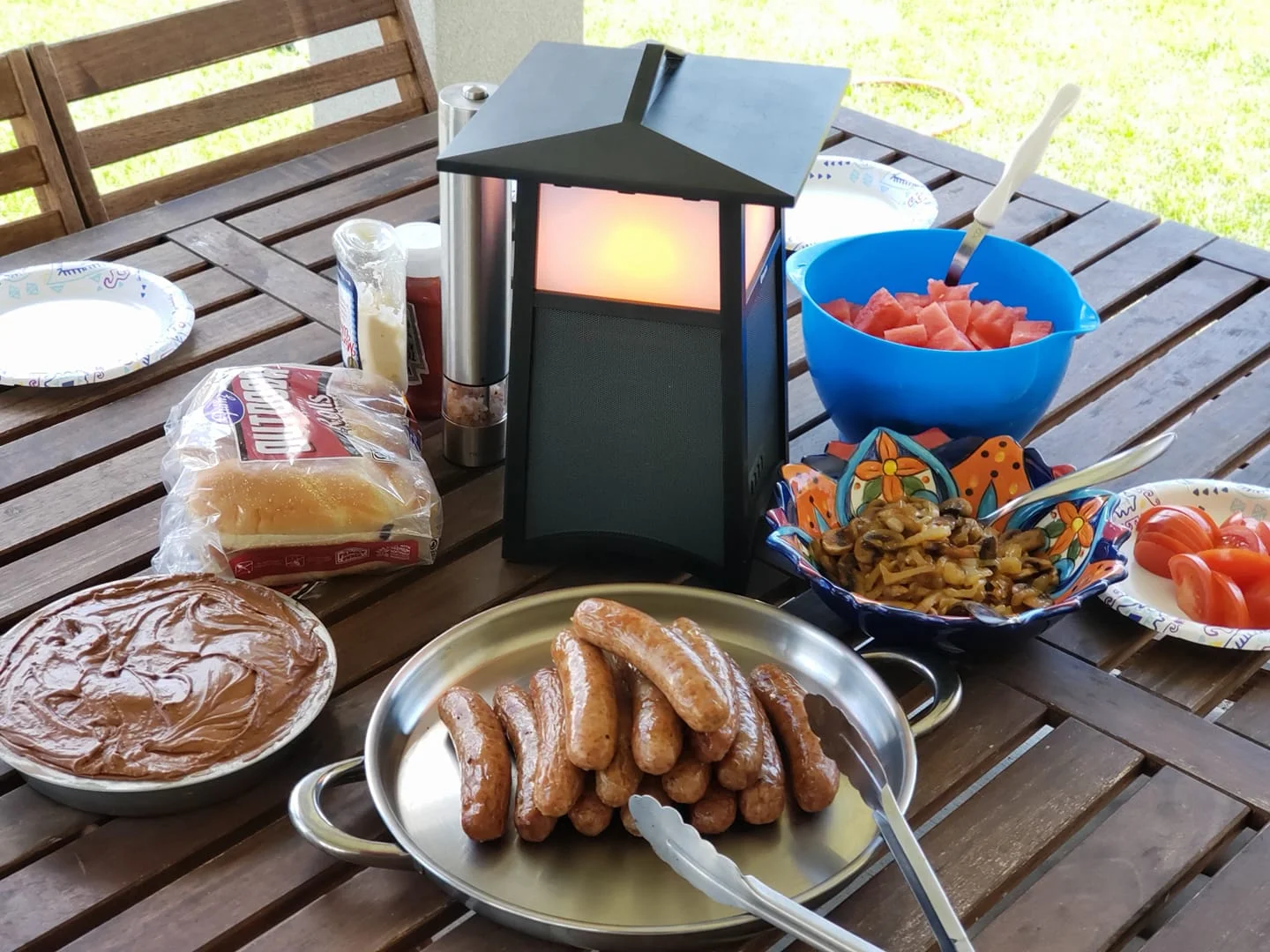 A picnic table set with food items including sausages, bread, and watermelon, accompanied by a lantern.
