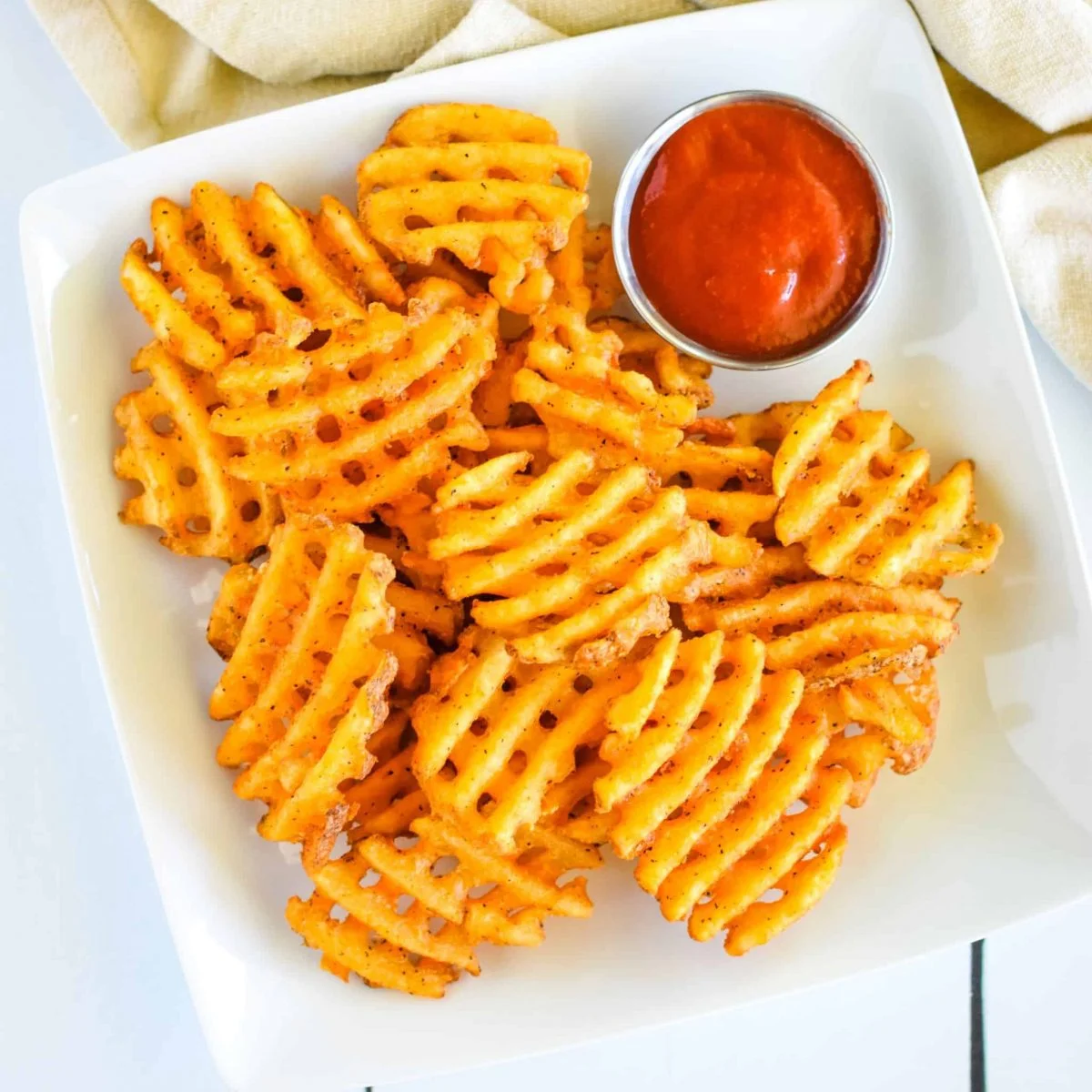 A serving of waffle fries arranged in a pile on a white plate, accompanied by a small cup of dipping sauce.