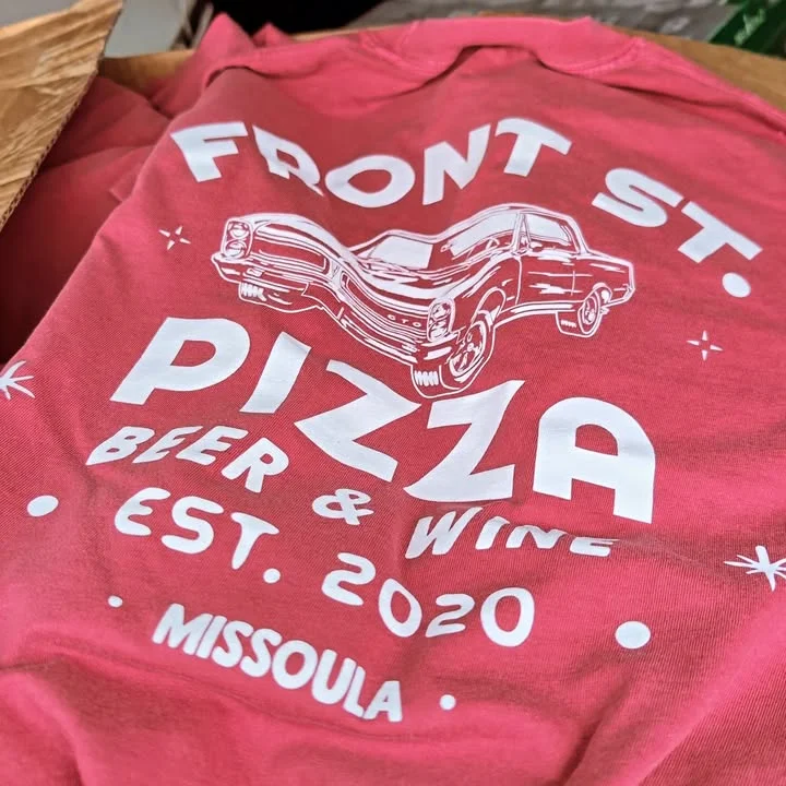 A close-up of a colorful t-shirt featuring a vintage car graphic with text about a pizza restaurant and its offerings.
