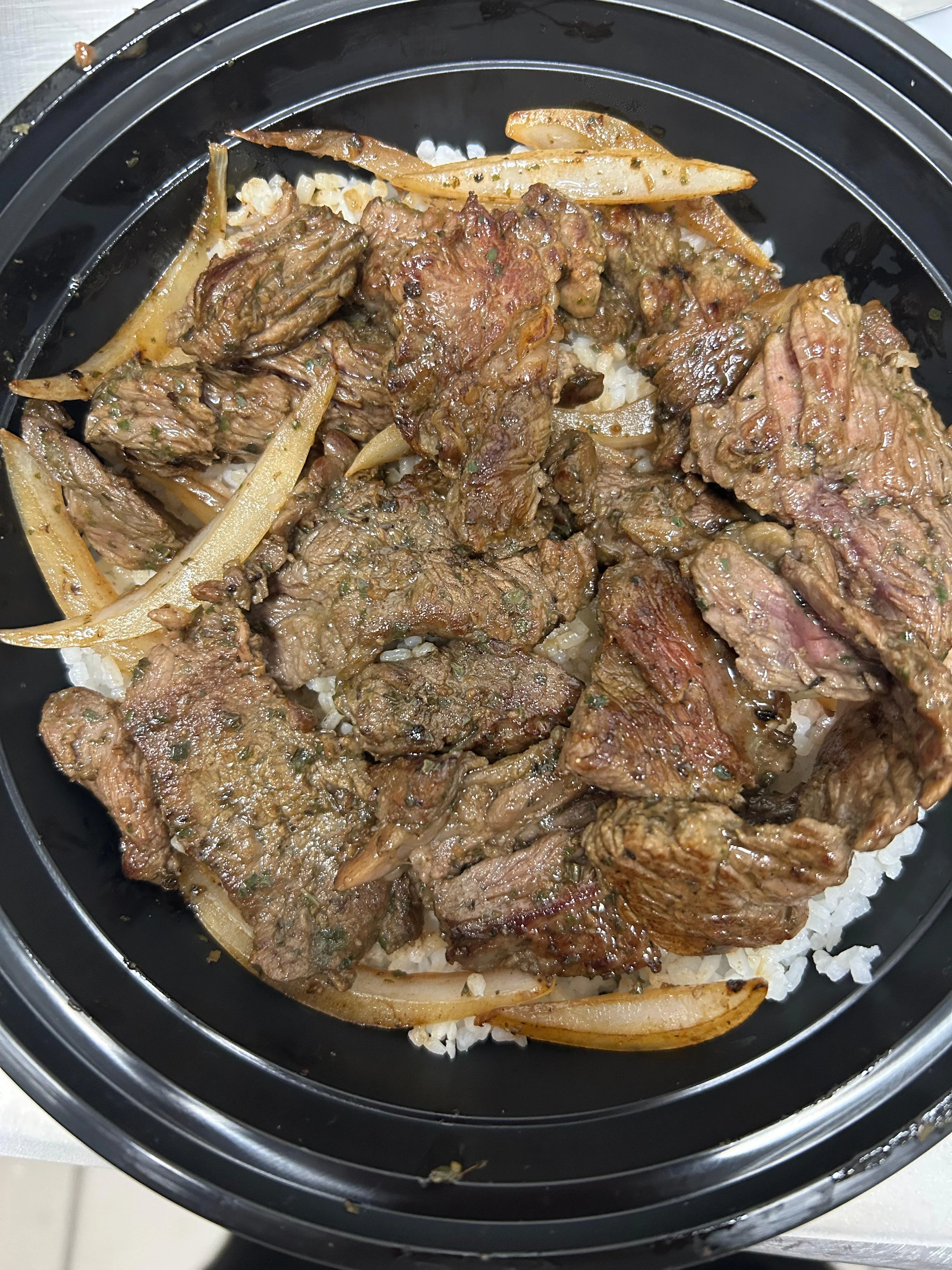 A takeout container filled with grilled beef strips over a bed of rice, accompanied by sliced onions, seasoned and perfectly cooked.