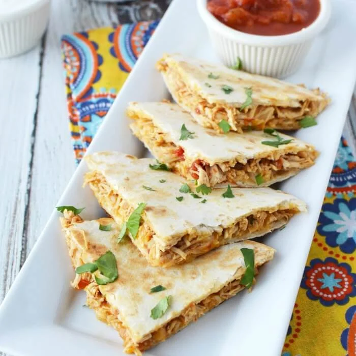 Three triangular slices of quesadilla served on a white plate, accompanied by a small bowl of salsa and garnished with fresh herbs.